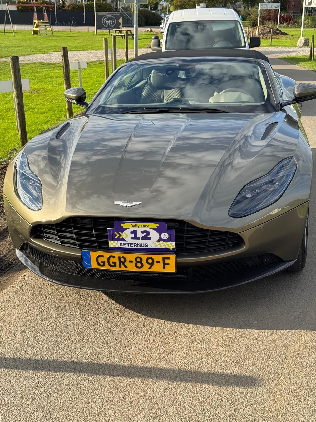 Een groene Aston Martin cabriolet met een Nederlands kenteken staat geparkeerd op een verharde parkeerplaats.
