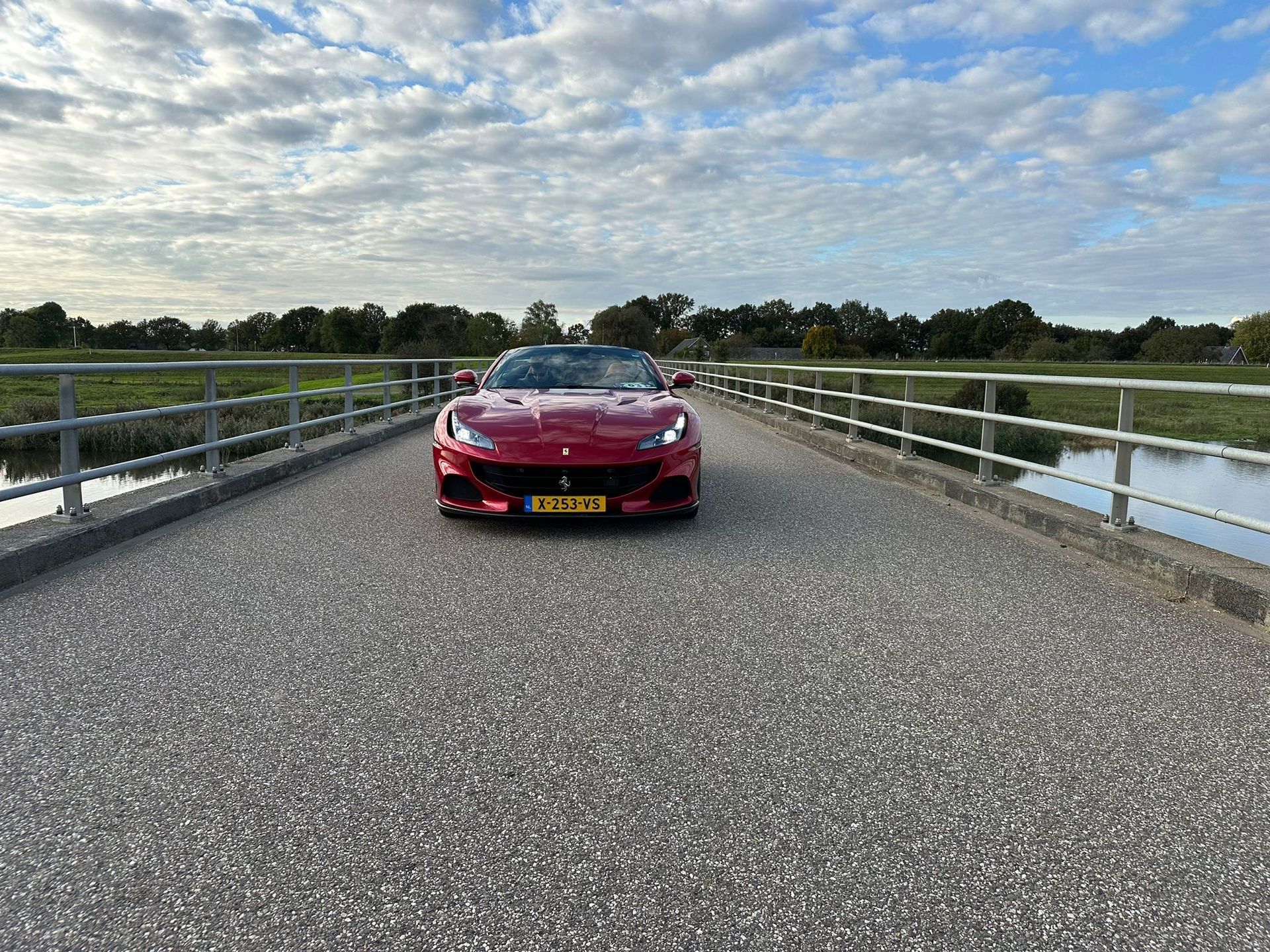 Rode Ferrari cabriolet op een brug, vooraanzicht. Bewolkte lucht, water aan de zijkanten, bomen op de achtergrond.