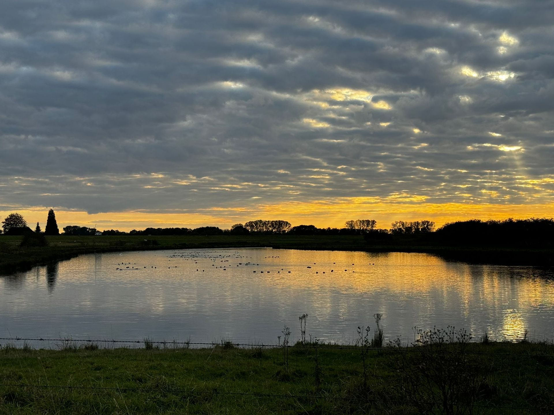 Zonsondergang boven een kalm meer, met donkere wolken en een geel-oranje lucht die in het water weerspiegeld wordt.