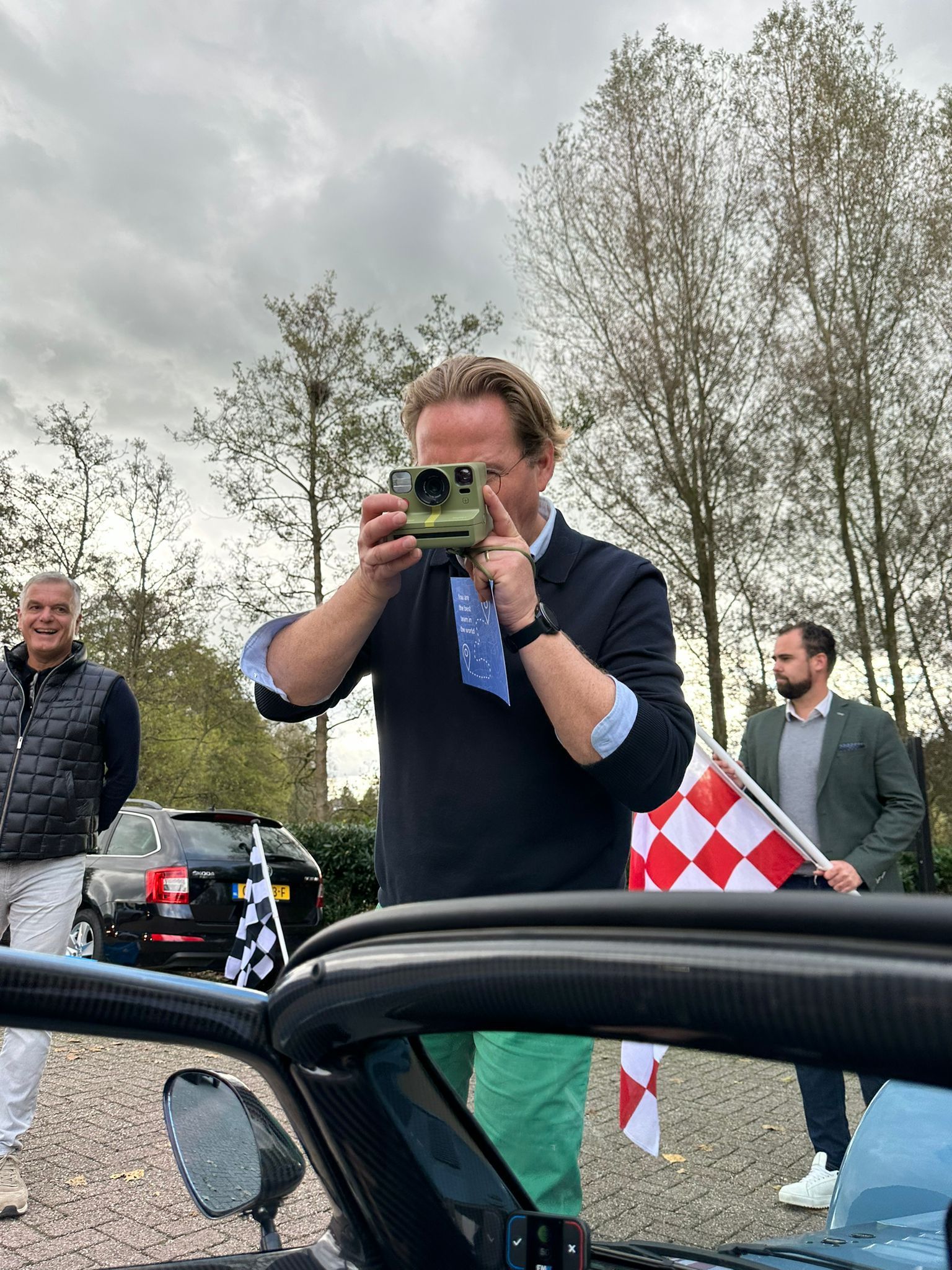 Een man in een blauw shirt en een groene broek maakt een foto naast een auto, terwijl hij een geblokte vlag vasthoudt. Twee andere mannen staan ​​in de buurt.