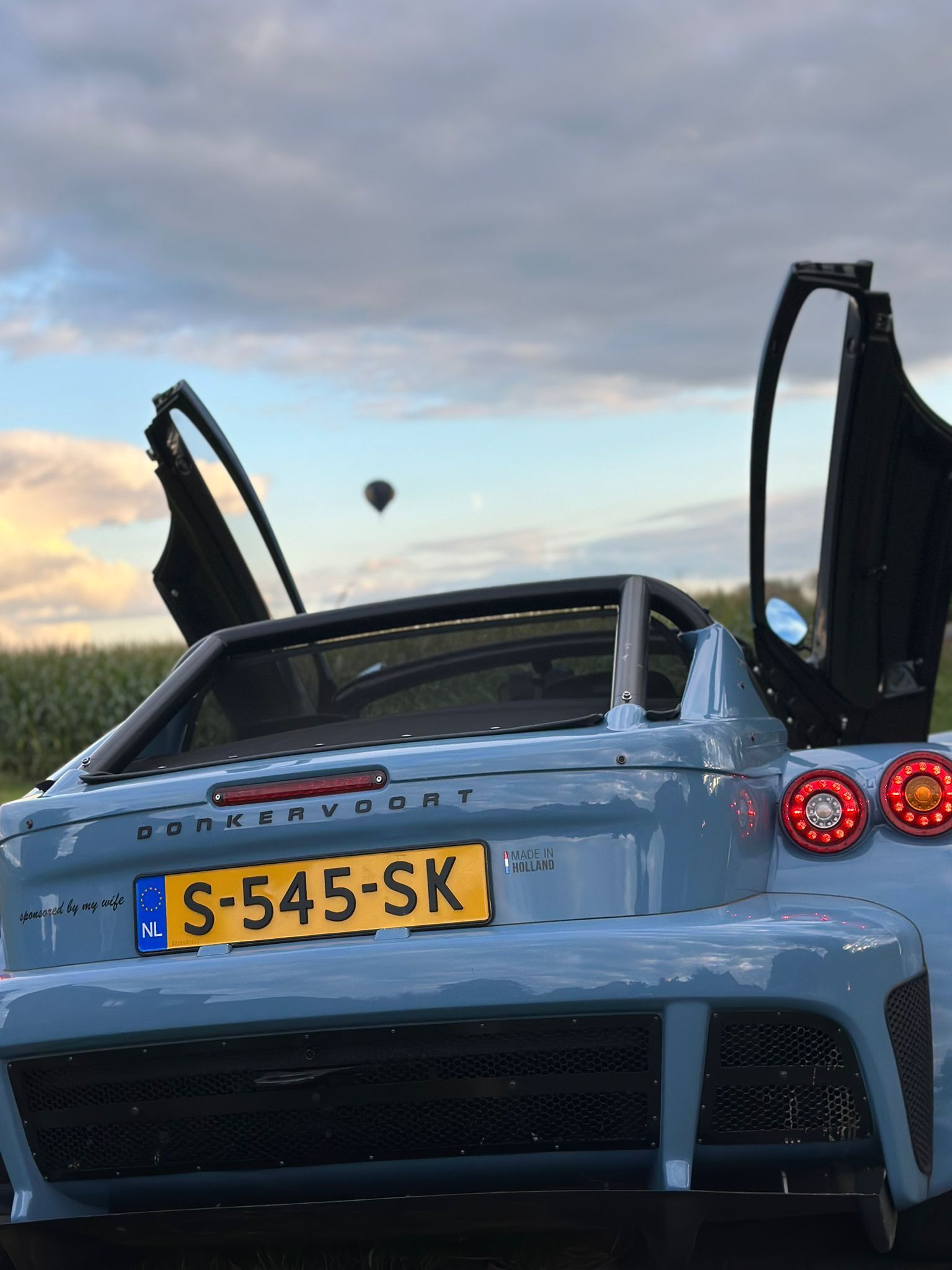 Blauwe sportwagen met open vleugeldeuren, met de rug naar de kijker toe. Kenteken S-545-SK. Heteluchtballon in de lucht.