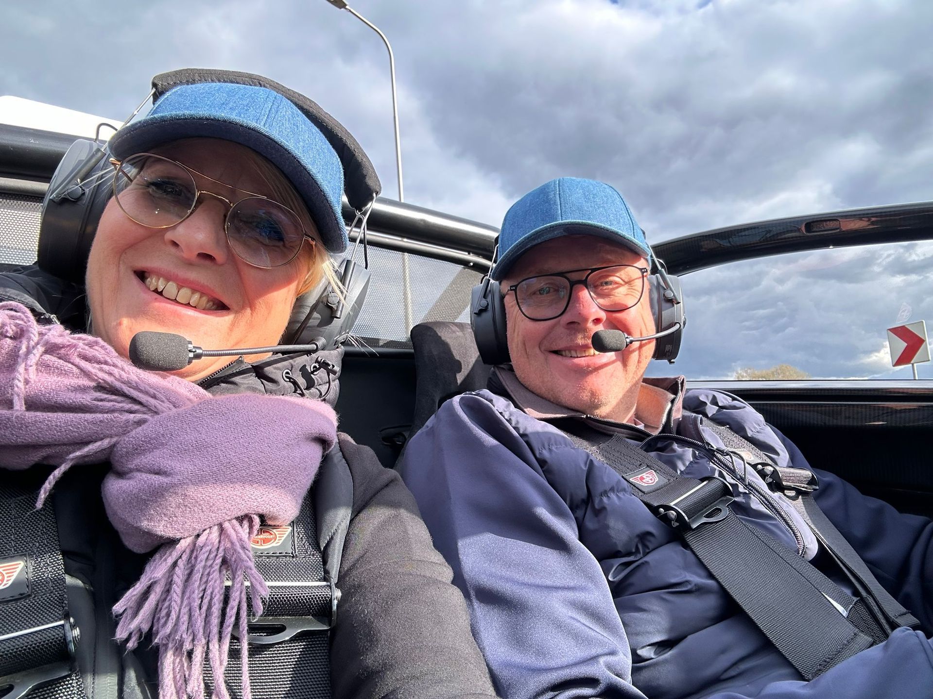 Twee mensen zitten lachend in de cockpit van een klein vliegtuig. Ze dragen headsets en petten tegen een bewolkte hemel.