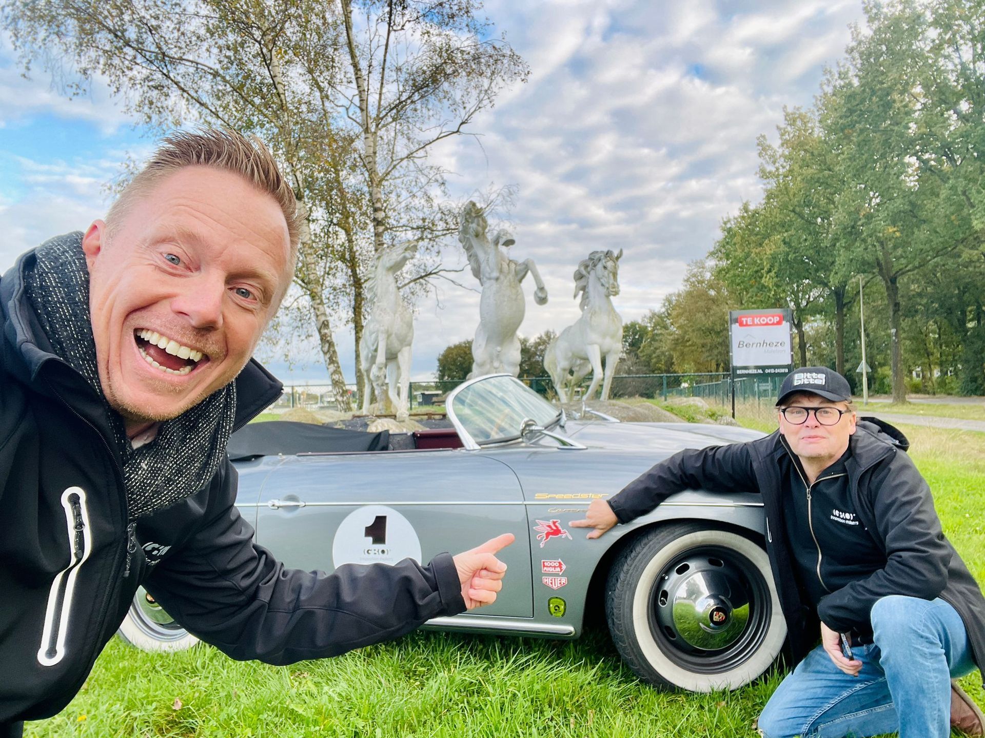 Twee mannen staan ​​bij een klassieke zilveren auto met paardenbeelden op de achtergrond. Een van de mannen wijst enthousiast.