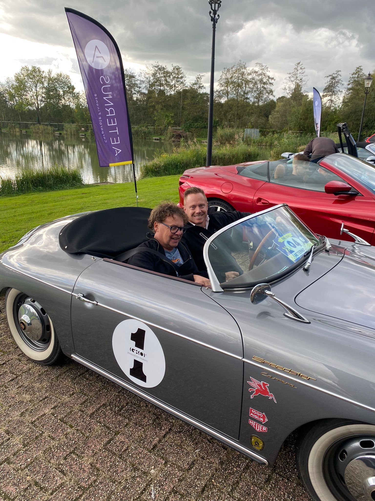 Twee mannen in een grijze cabriolet met een wit nummer één, geparkeerd bij een meer.