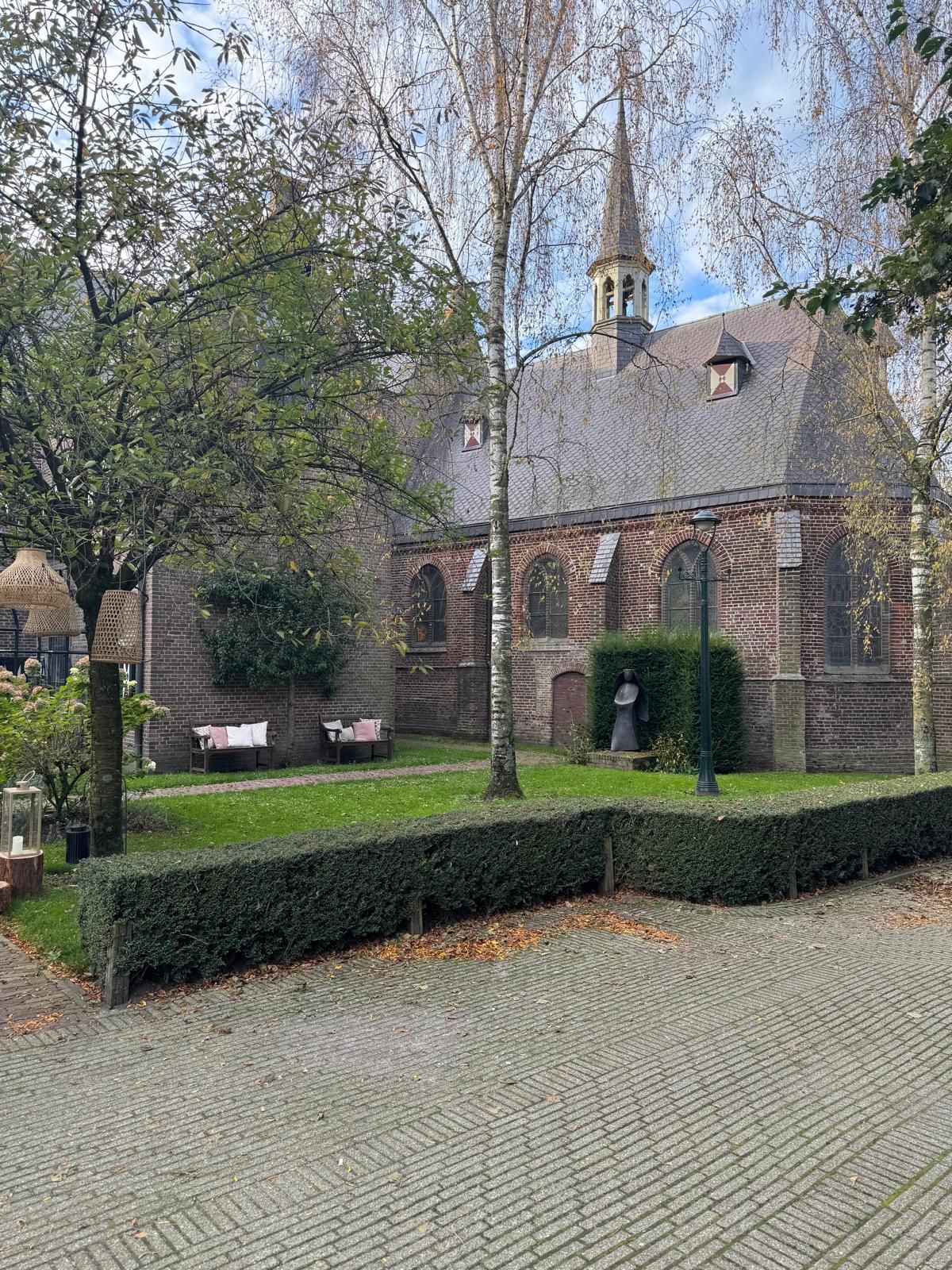 Oude bakstenen kerk met een torenspits, omgeven door bomen en een keurig onderhouden gazon.