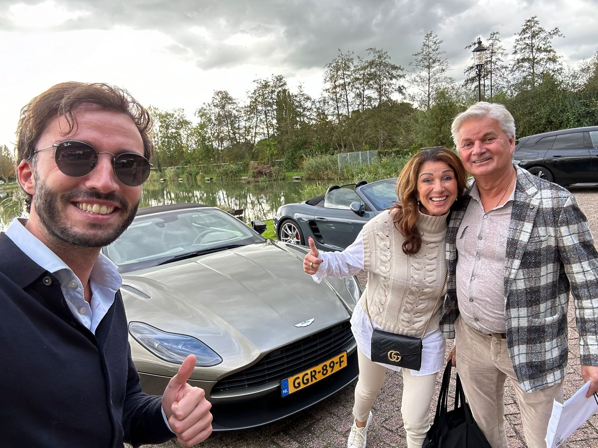 Drie mensen poseren bij een zilverkleurige sportwagen vlakbij het water. Een man met een zonnebril steekt zijn duim omhoog. Een vrouw maakt een gebaar.