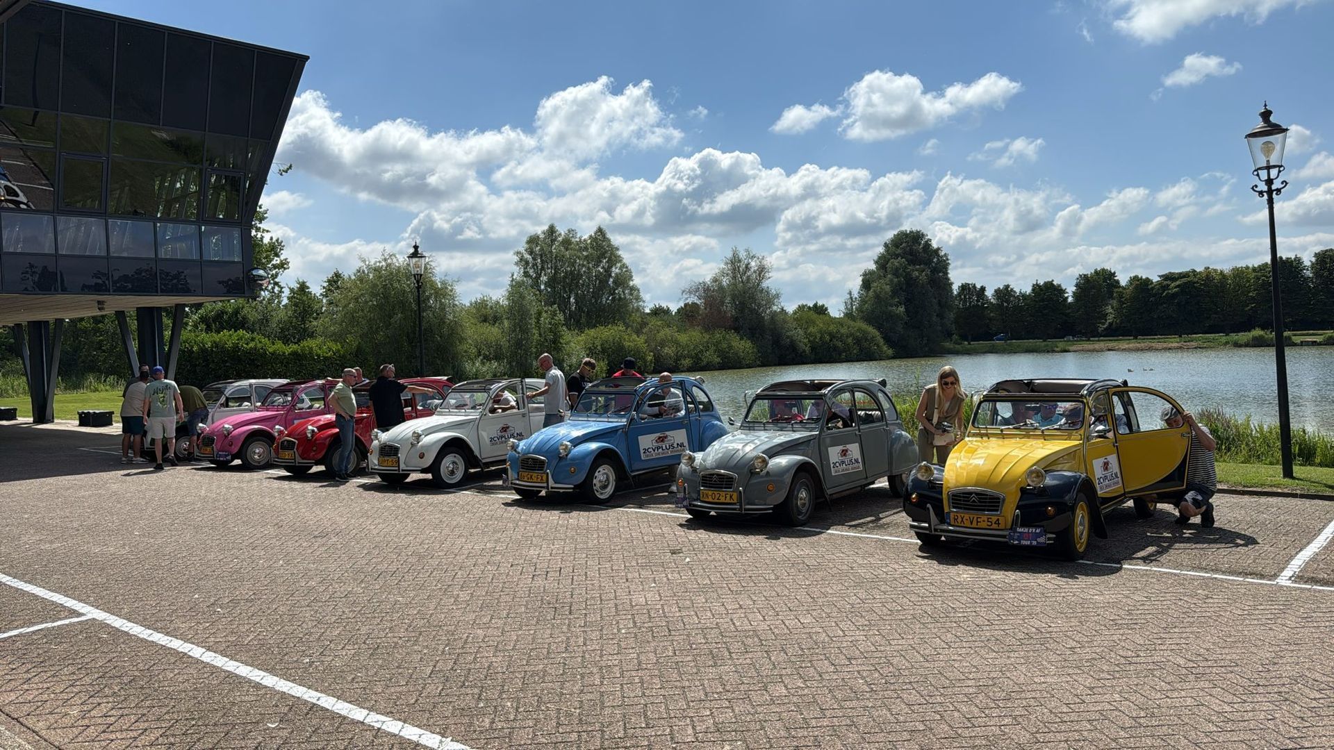 Klassieke auto's geparkeerd op een stenen ondergrond, naast een meer en een gebouw. ​​Zonnige, blauwe lucht.