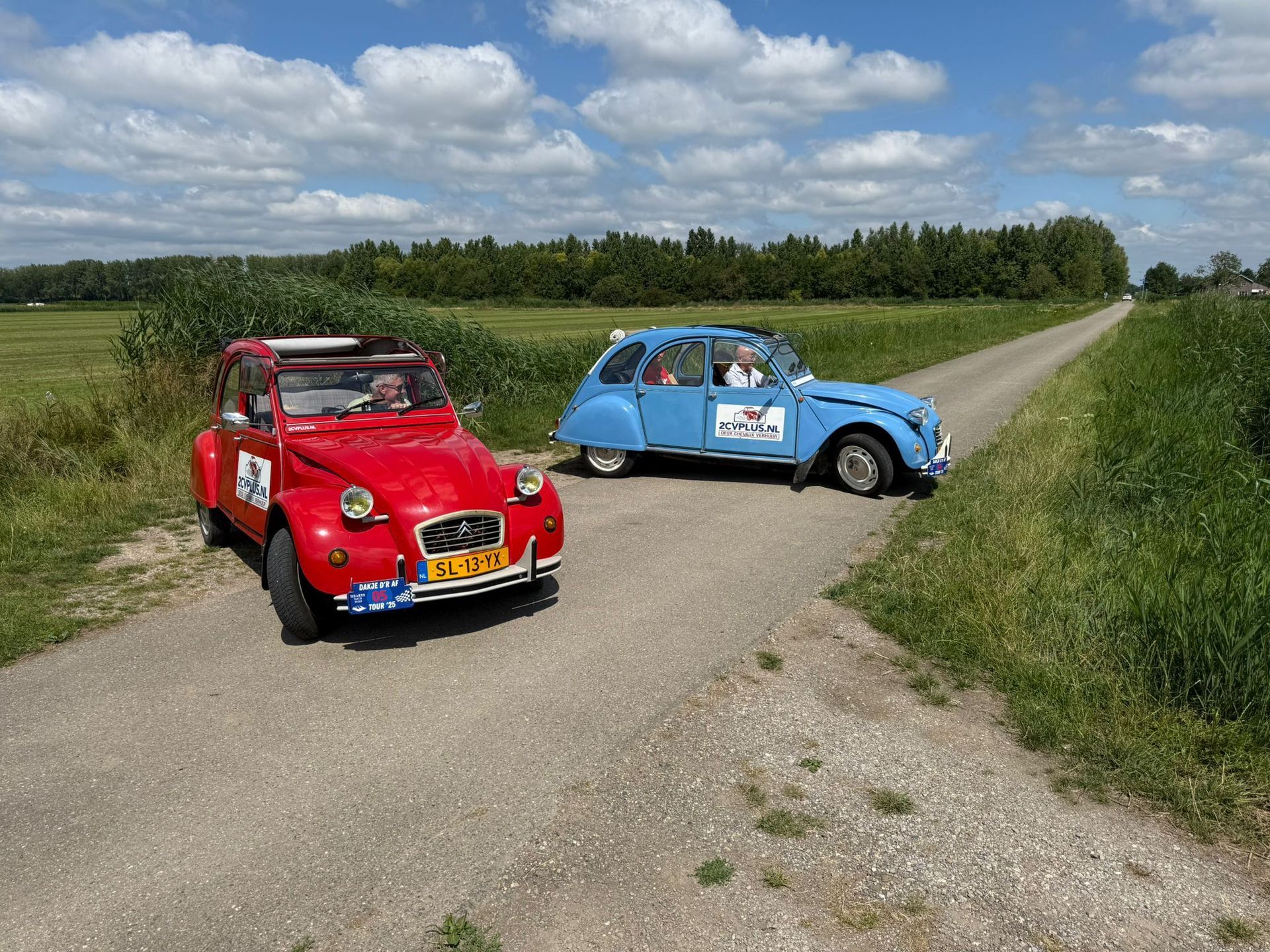Twee klassieke Citroën 2CV's: een rode en een blauwe, op een grindweg naast een veld. Zonnige dag.