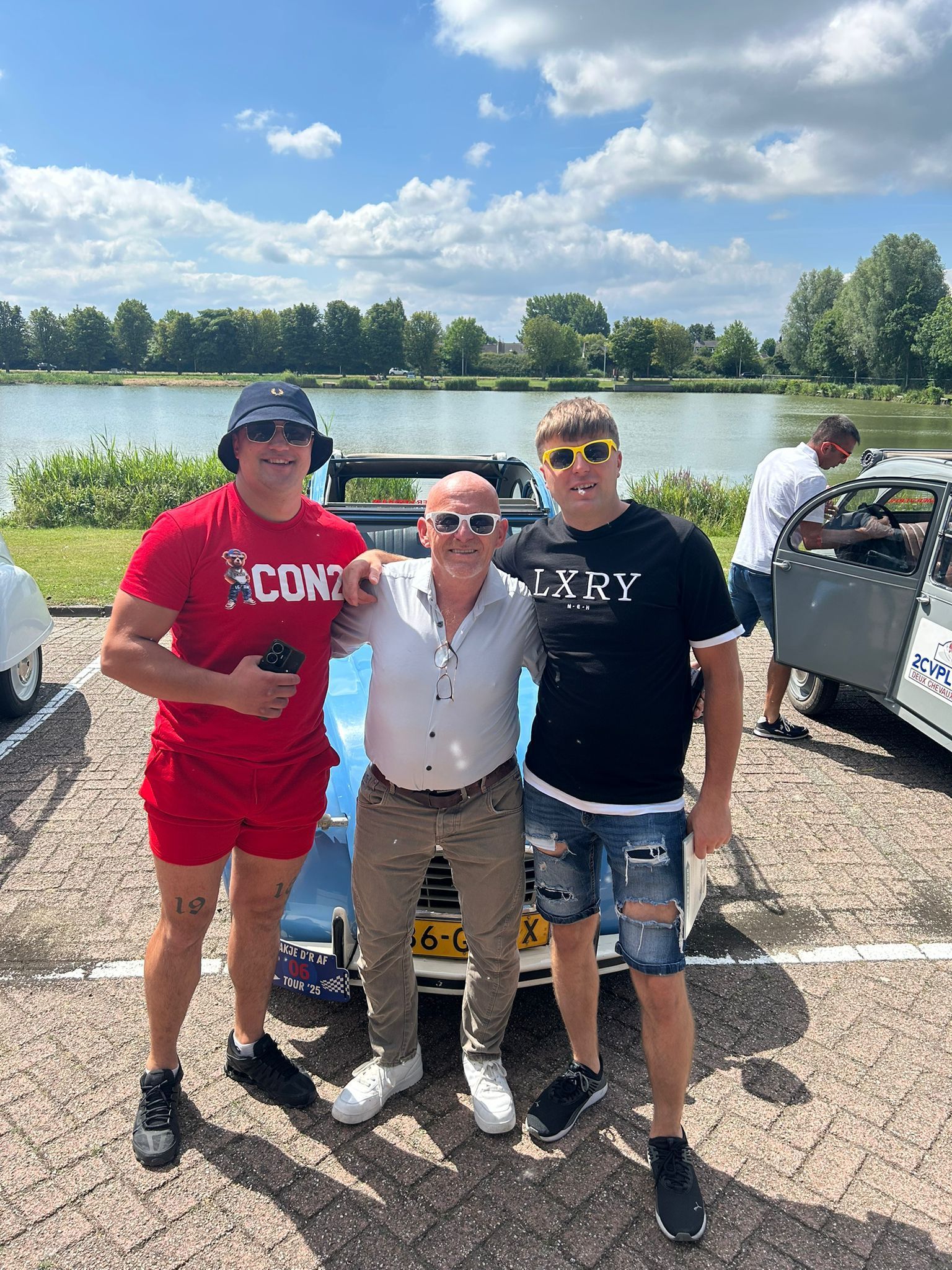 Drie mannen poseren voor een blauwe cabriolet, met een meer op de achtergrond. Zonnige dag.