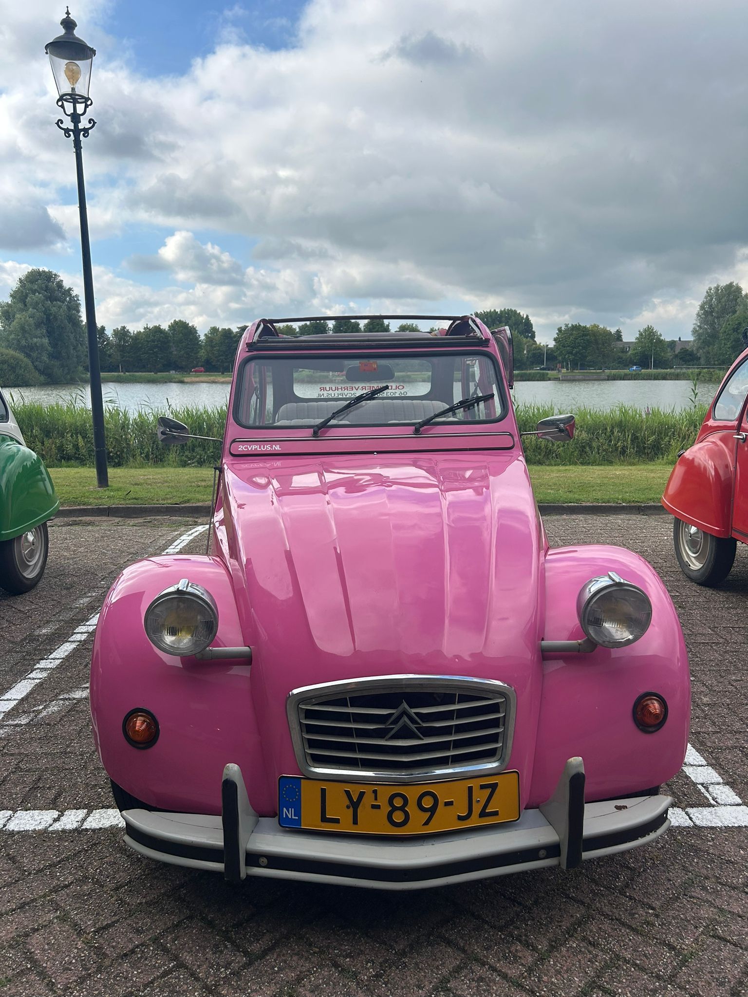 Een roze Citroën 2CV met Nederlands kenteken staat geparkeerd op een parkeerplaats. Aan weerszijden is nog een auto gedeeltelijk zichtbaar.