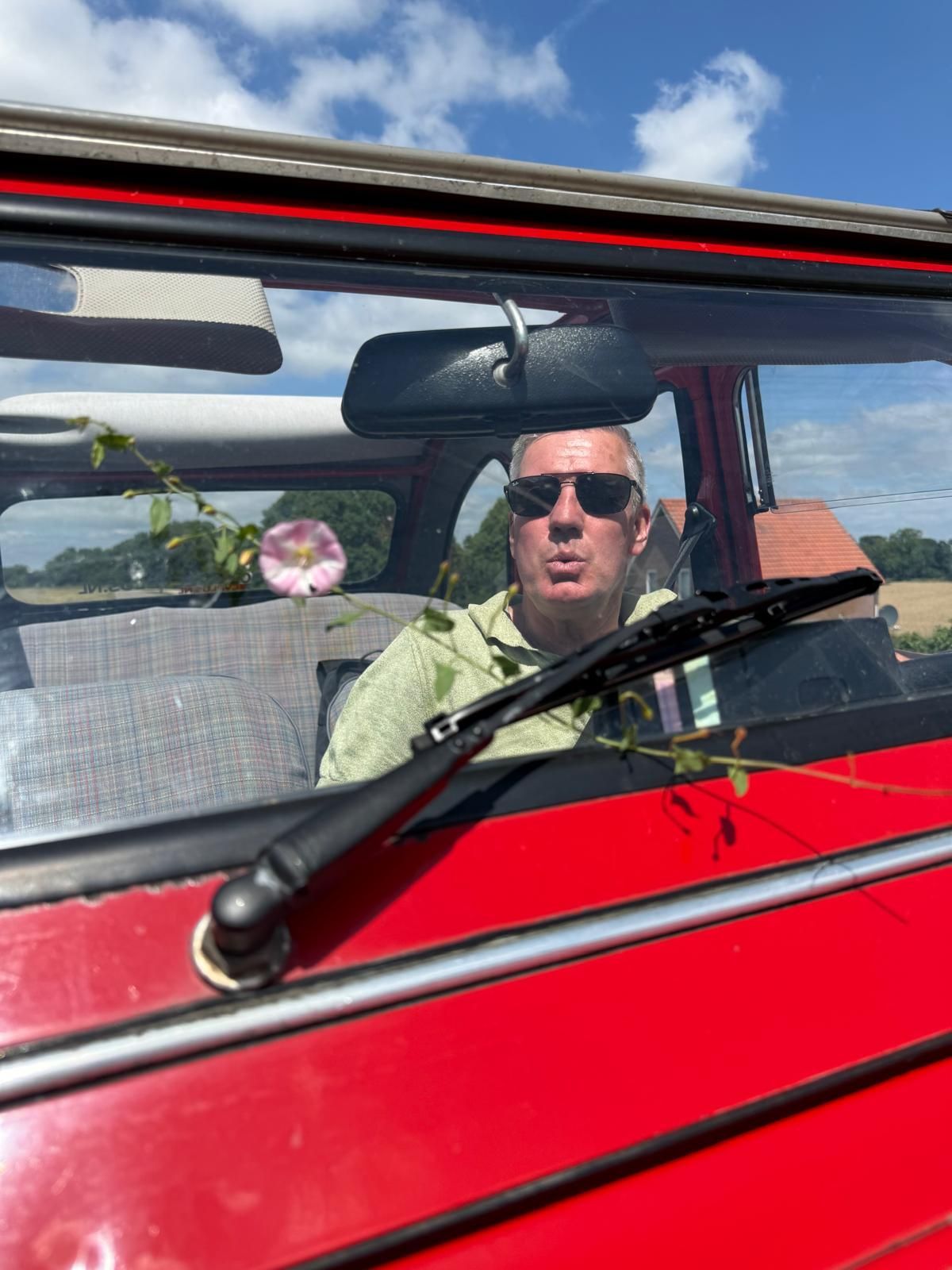 Een man met een zonnebril zit in een rode klassieke auto, die door de voorruit te zien is.