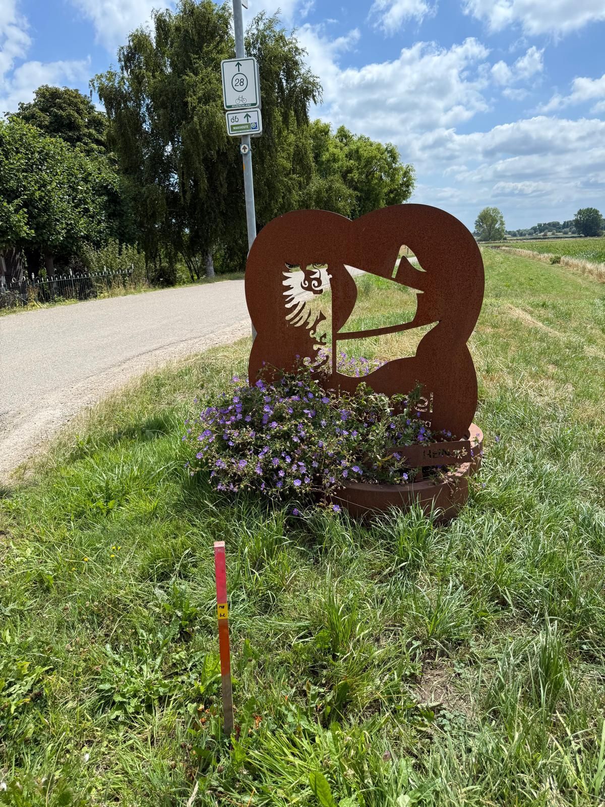 Metalen hartsculptuur met bloemen naast een pad en een wegwijzer, op een grashelling.