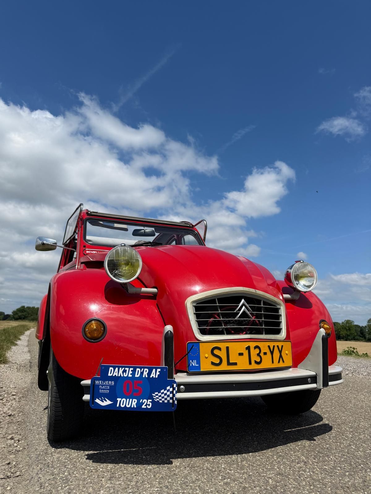 Een rode oldtimer Citroën 2CV staat geparkeerd op een onverharde weg, met een blauwe lucht op de achtergrond.