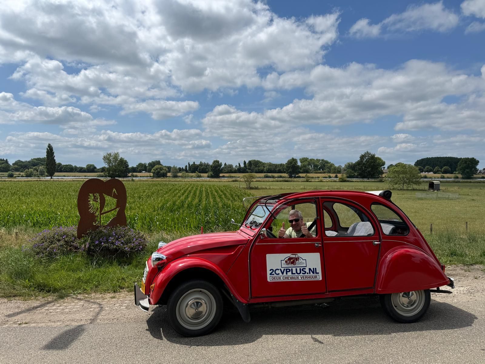 Een rode Citroën 2CV op een weg in een landelijke omgeving, met een persoon achter het stuur.