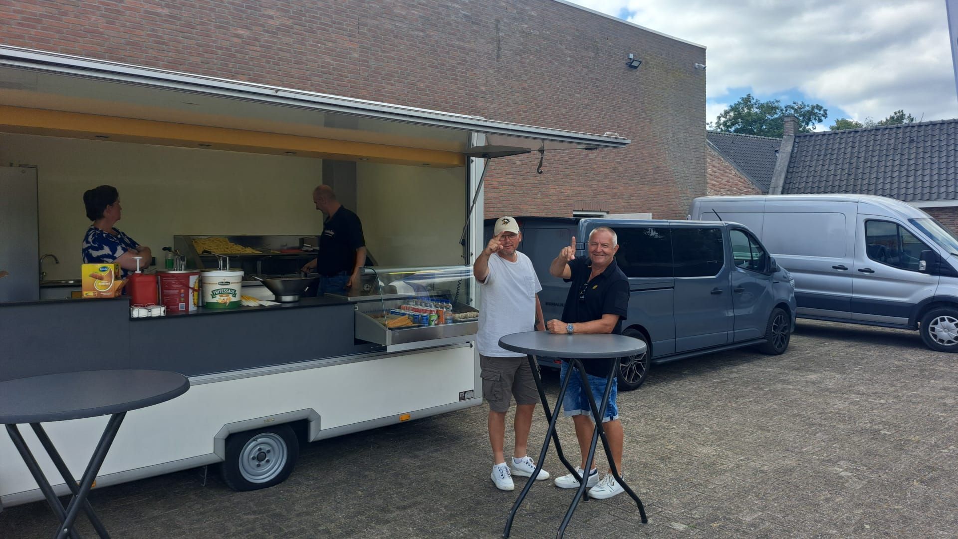 Twee mannen steken hun duim omhoog bij een foodtruck met twee medewerkers. Er staan ​​busjes in de buurt geparkeerd.