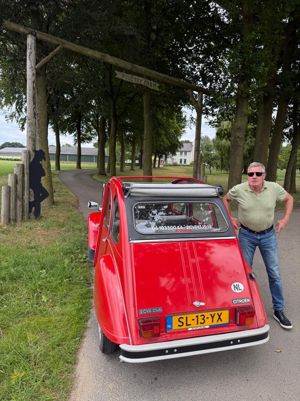 Een rode oldtimer staat geparkeerd onder een houten poort, met een man ernaast.