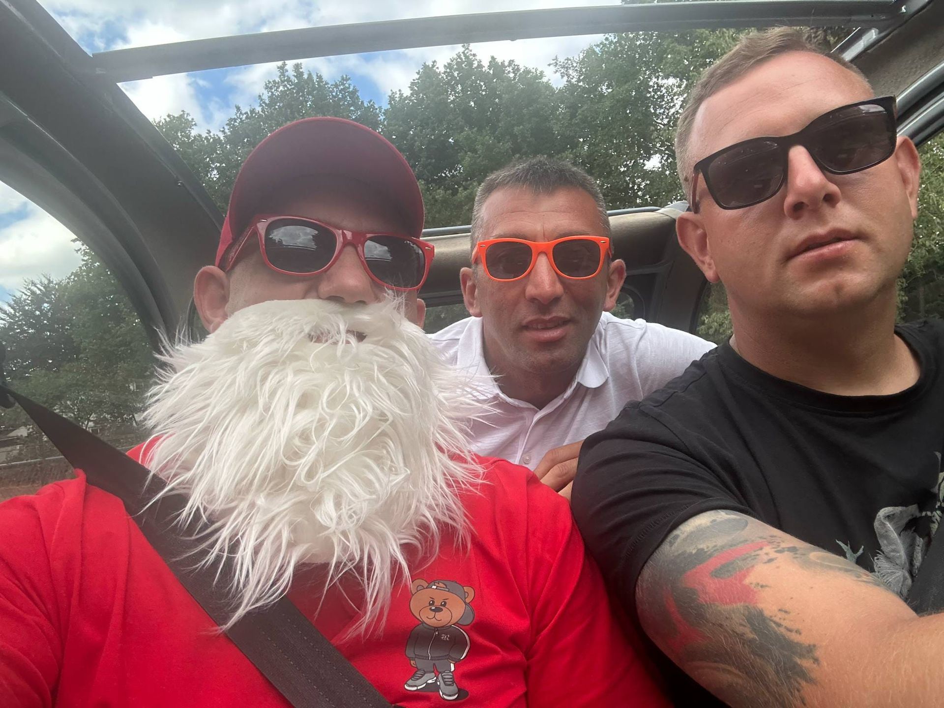 Drie mannen in een voertuig, één met een kerstmanbaard en een rood shirt, allen met een zonnebril op.