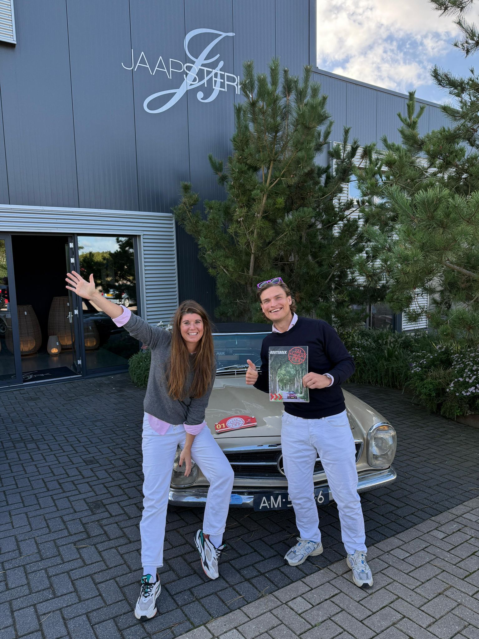 Twee mensen poseren voor een gebouw met een auto. De vrouw zwaait, de man houdt een tijdschrift vast.