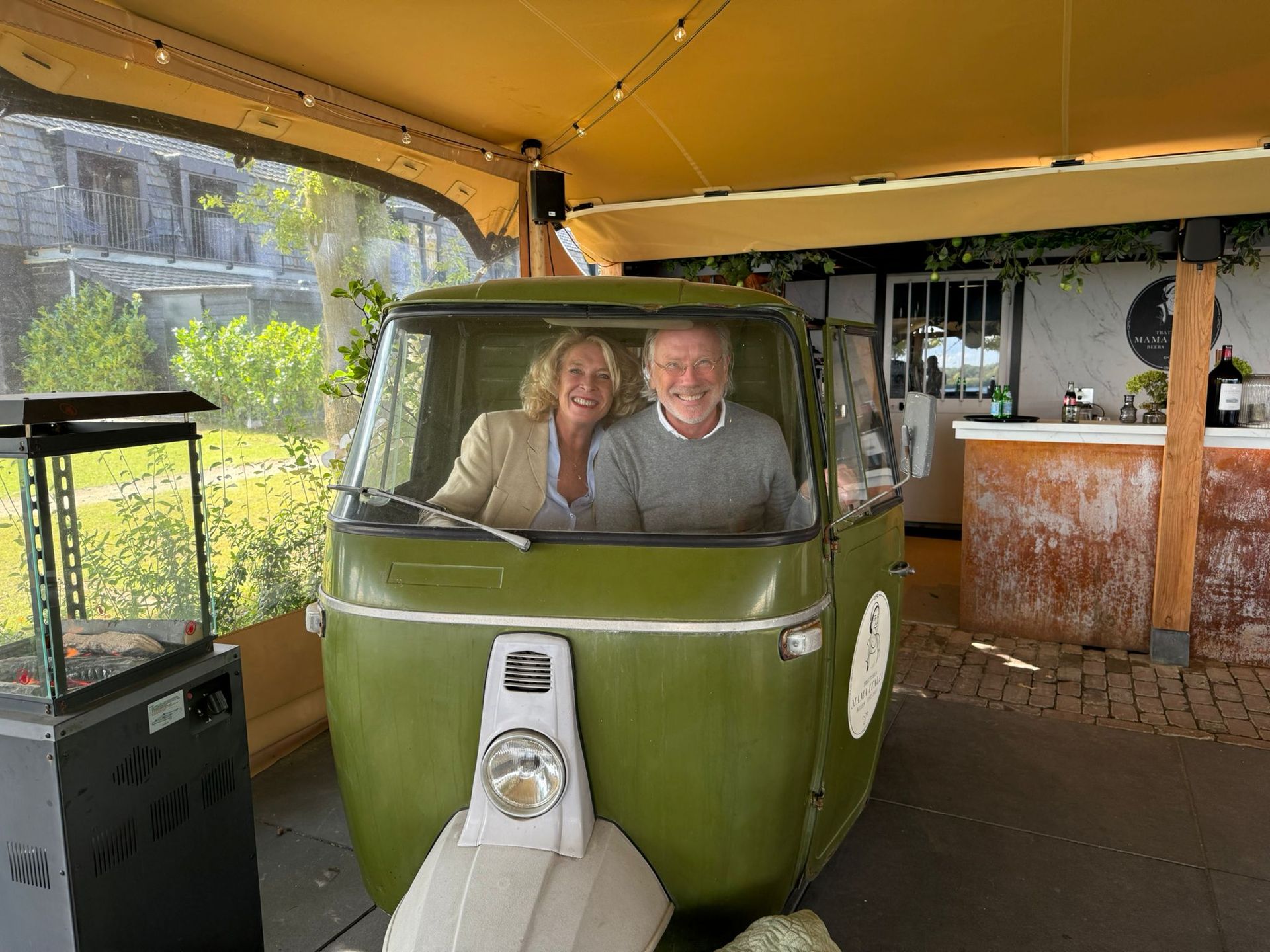 Een stel in een groene driewieler, onder een beige luifel; op de achtergrond is een bar te zien.