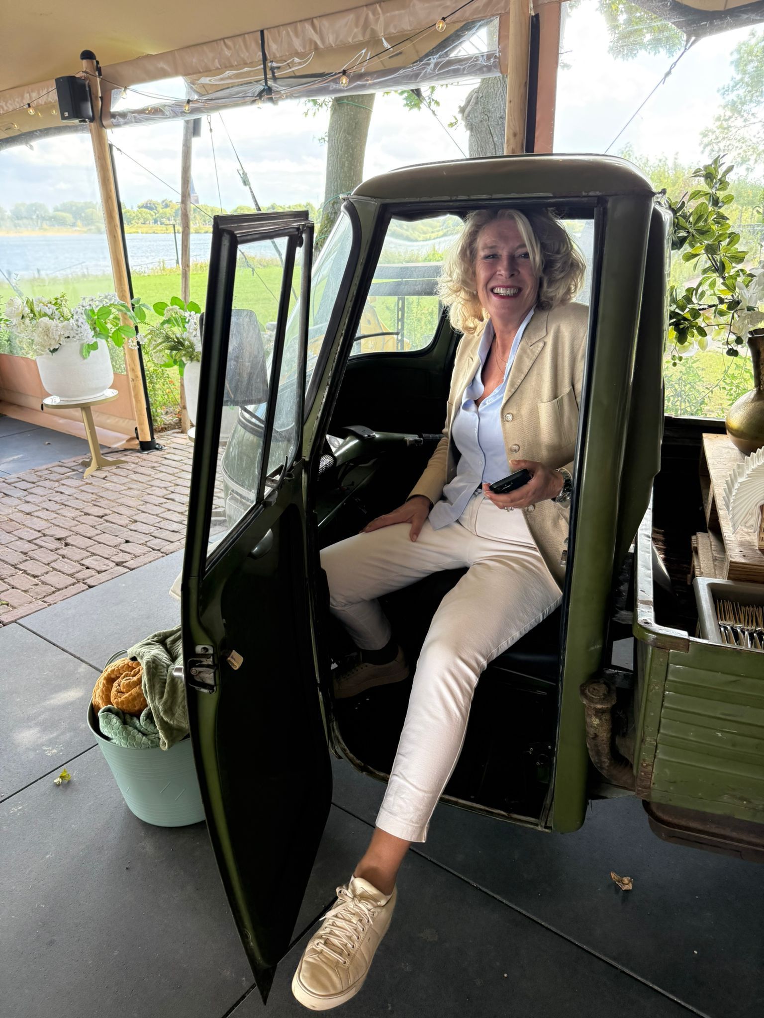 Vrouw zit in een groene auto met open deur en lacht. Lichtbeige jasje, witte broek. Buitenomgeving.