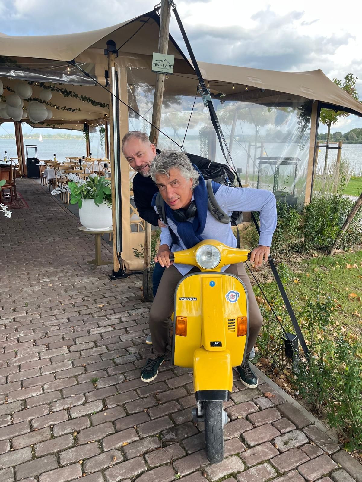 Twee mannen op een felgele scooter. Eén man bestuurt de scooter en lacht. De ander rijdt achter hem. Een terrasje speelt zich af op een terras.