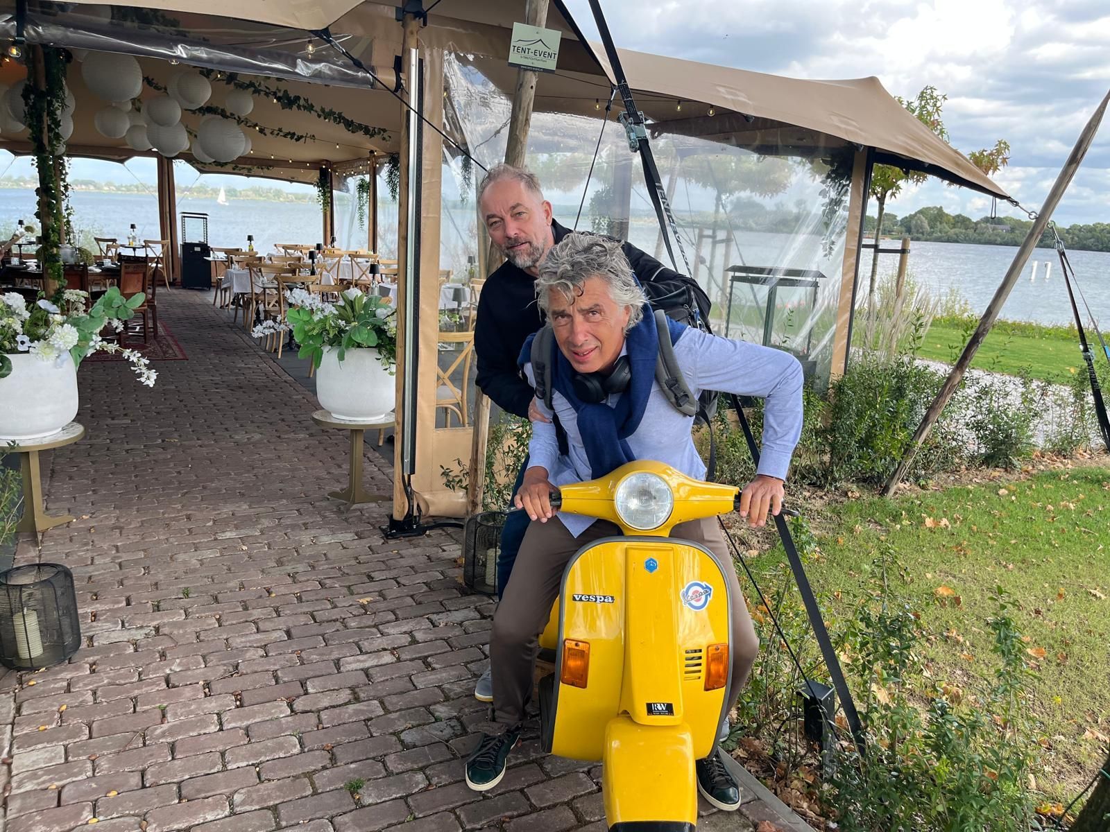 Twee mannen poseren speels op een gele Vespa-scooter bij een meer en een terrasrestaurant.