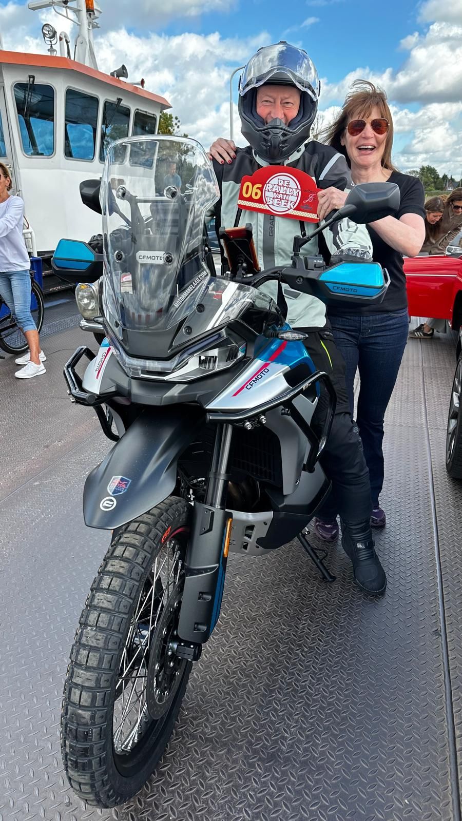 Man en vrouw met een motor op een veerboot. De man met helm houdt een bord vast; beiden glimlachen.