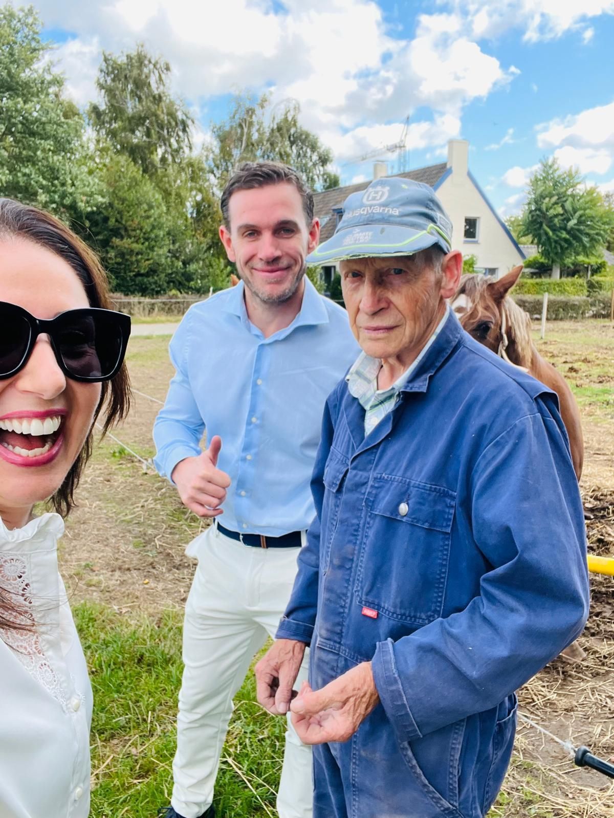 Drie mensen glimlachen buiten bij een paard. Een man in een blauw overall staat naast een man in een lichtblauw shirt. Een vrouw met een zonnebril maakt de foto.