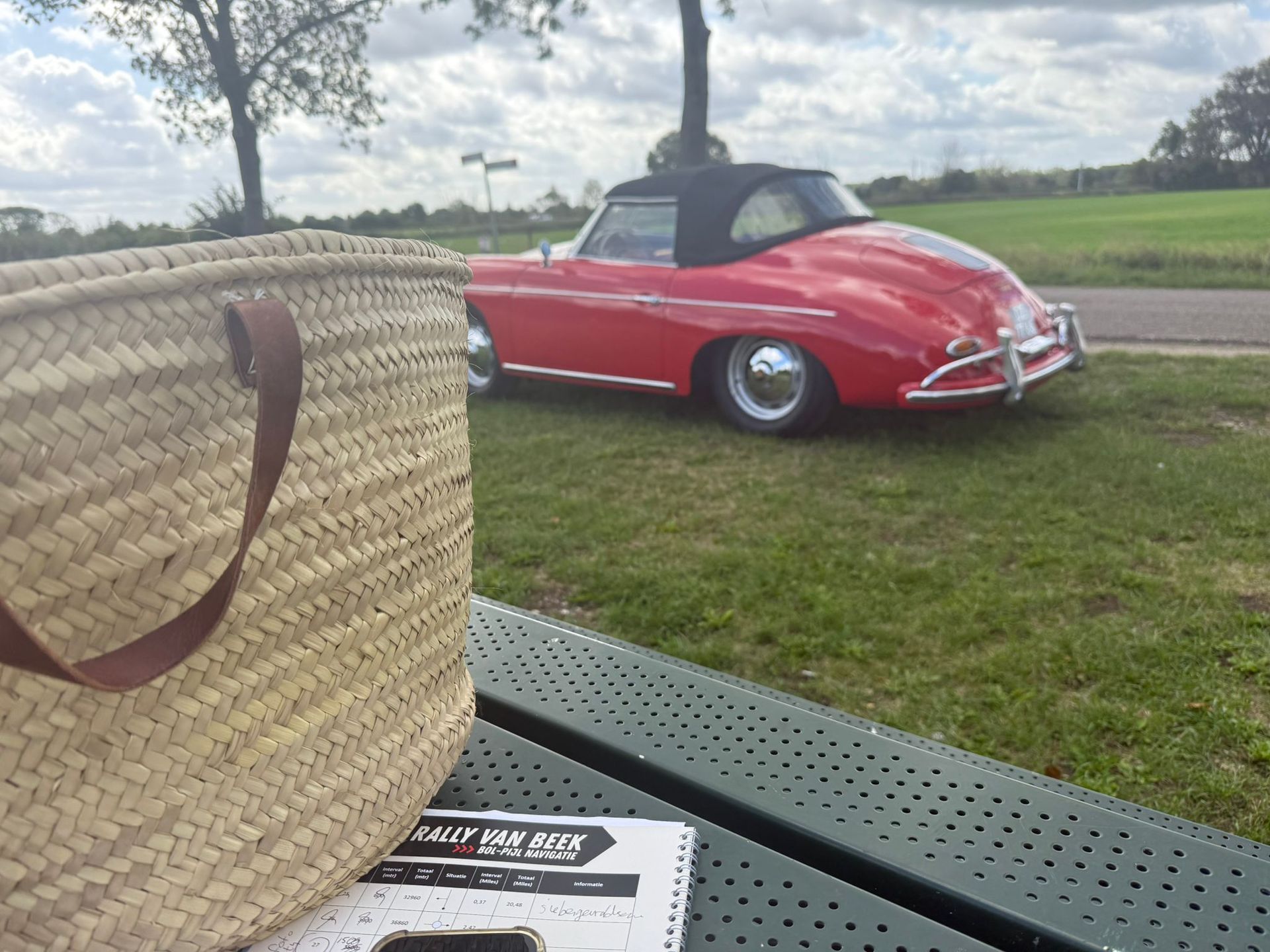 Een rode cabriolet staat geparkeerd op het gras naast een picknicktafel met een gevlochten tas en een notitieboekje.