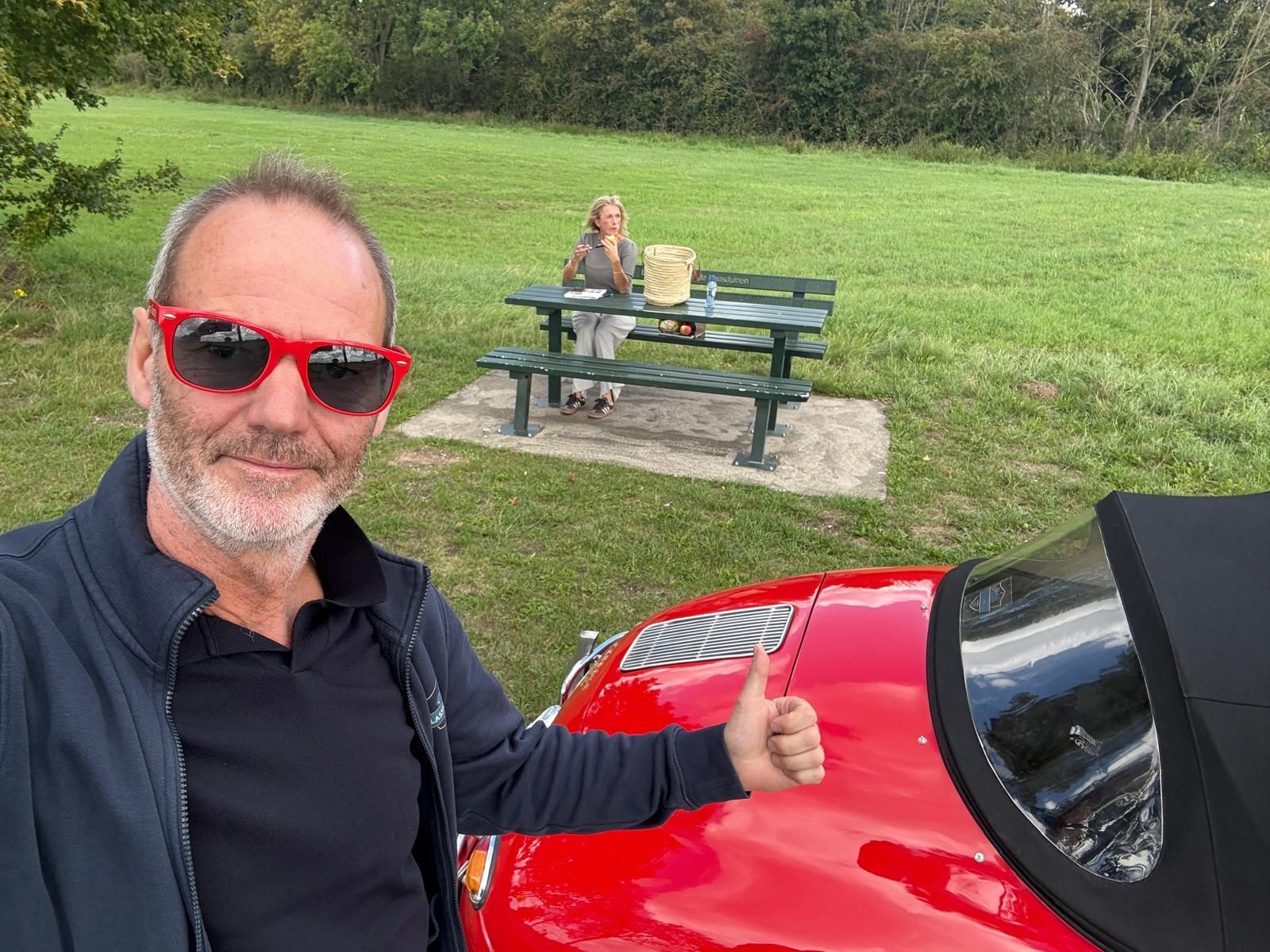 Een man met een rode zonnebril en een rode auto steekt zijn duim omhoog. Iemand zit aan een picknicktafel in een grasveld.