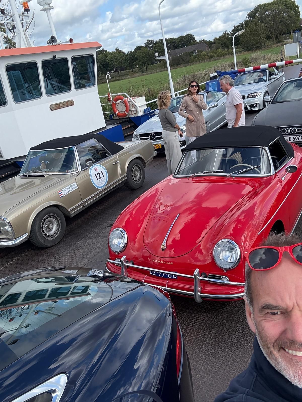 Een rode Porsche cabriolet en een beige auto op een veerboot, mensen staan eromheen. Een man maakt een selfie.