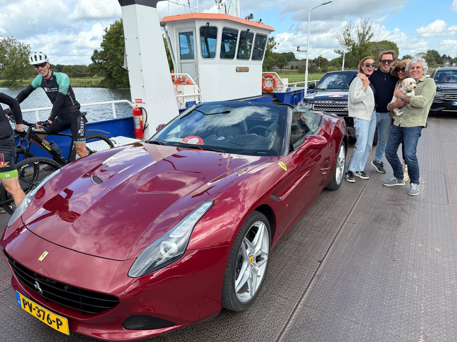 Een rode Ferrari cabriolet op een veerboot met een groep mensen, van wie één een hond vasthield, en een fietser.