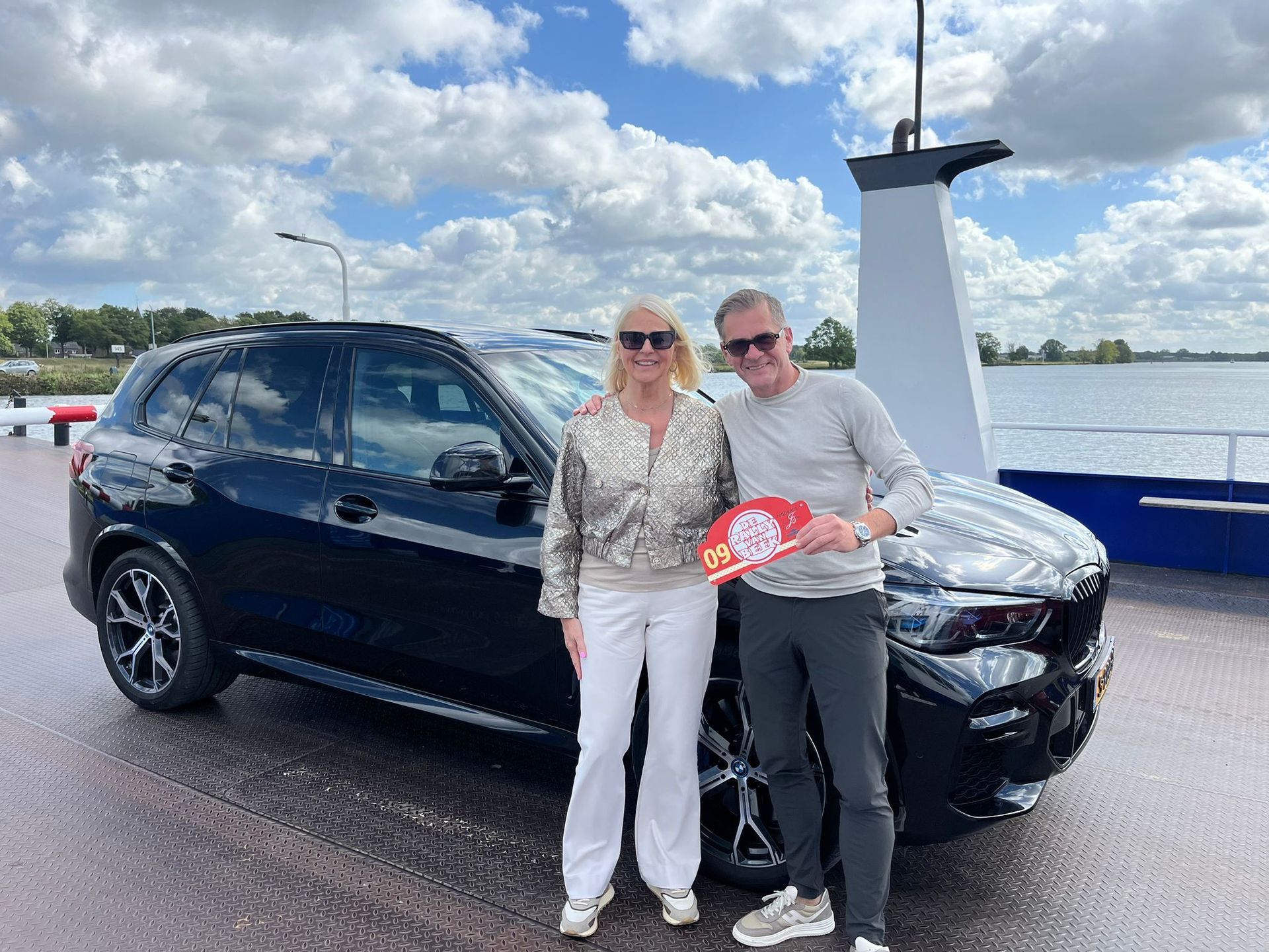Een stel staat naast een zwarte SUV op het dek van een veerboot; de man houdt een prijs vast; zonnig weer.
