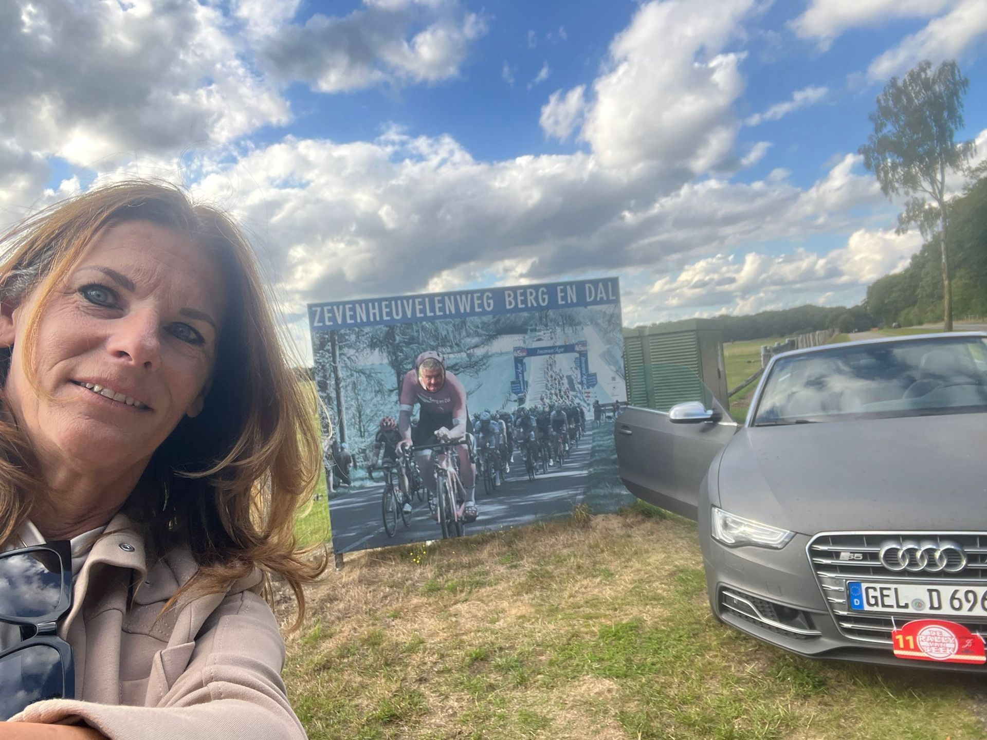 Vrouw maakt een selfie bij een bord met fietsers en een grijze auto onder een bewolkte hemel.