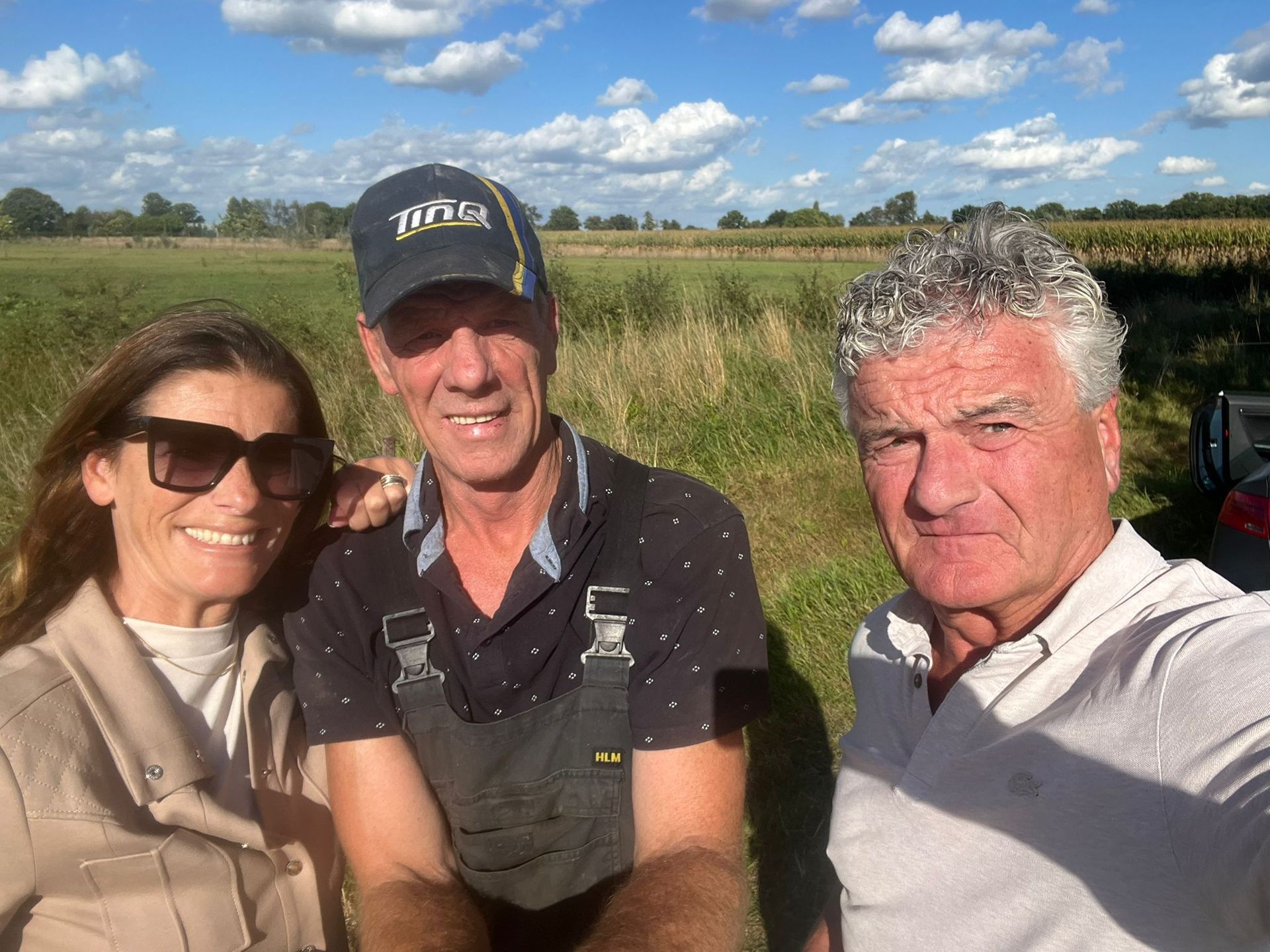 Drie mensen glimlachen buiten: een vrouw met een zonnebril, een man in een overall en een man in een wit shirt; een veld op de achtergrond.