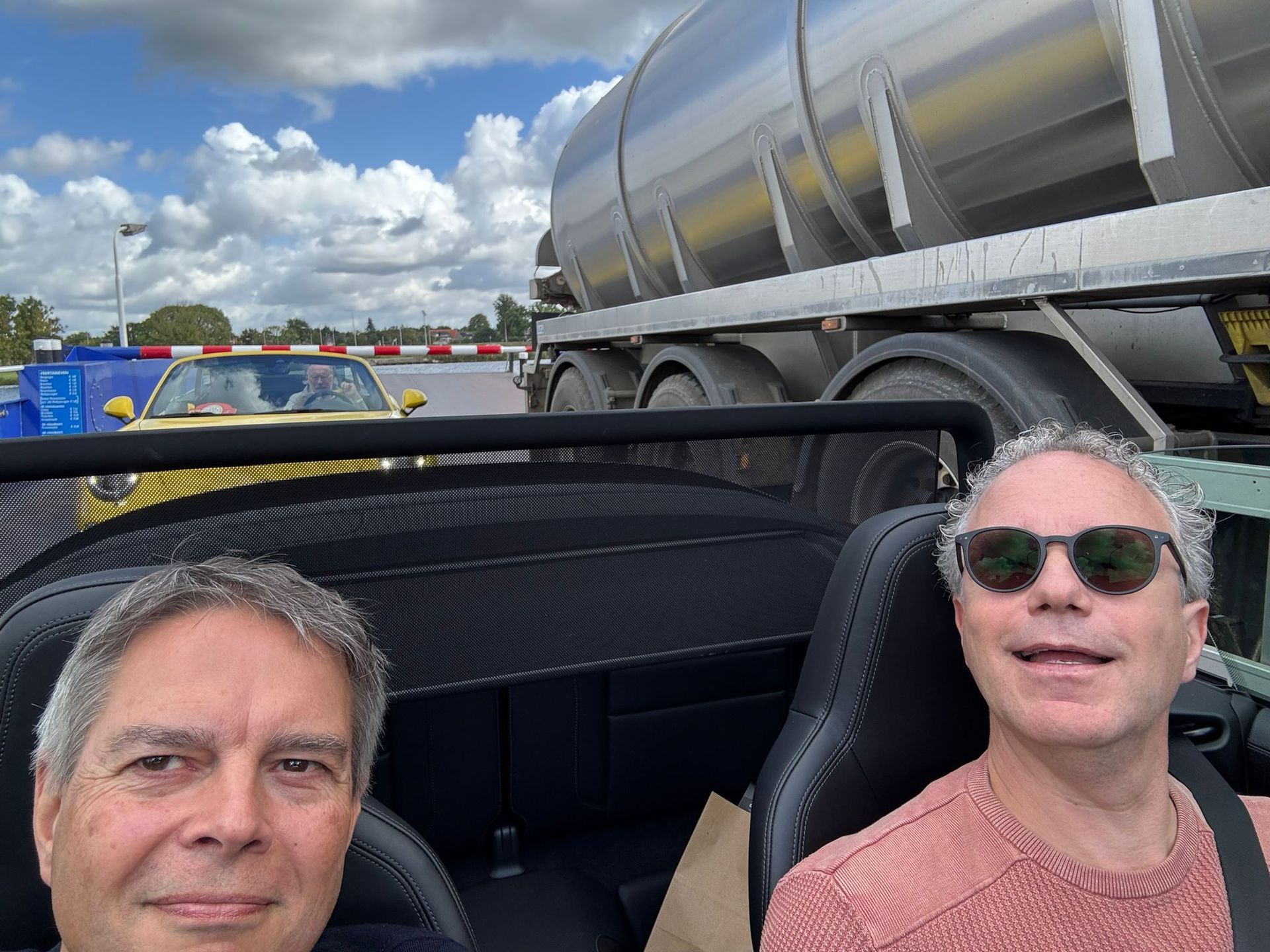 Twee mannen in een cabriolet, lachend. Achter hen een grote tankwagen. Zonnige, blauwe lucht.