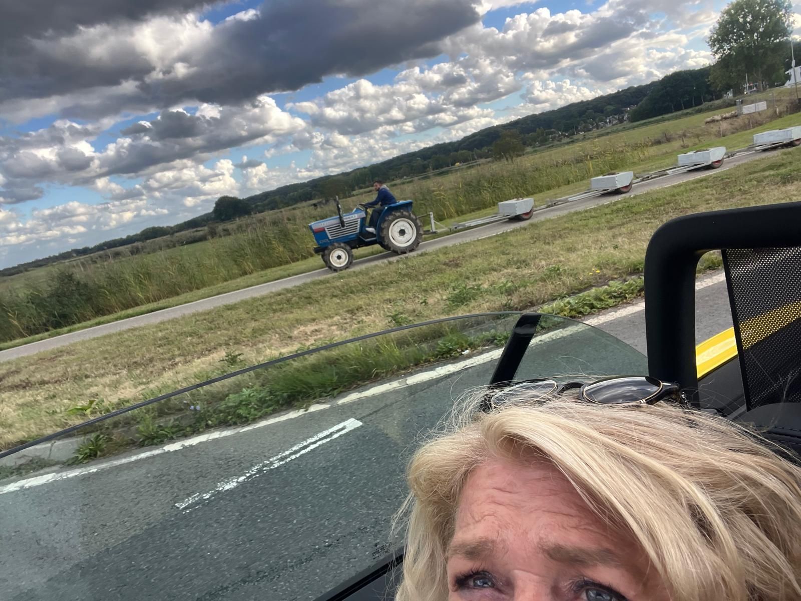 Vrouw maakt een selfie met een tractor die karren trekt op een weg, buiten. Bewolkte lucht, groen.
