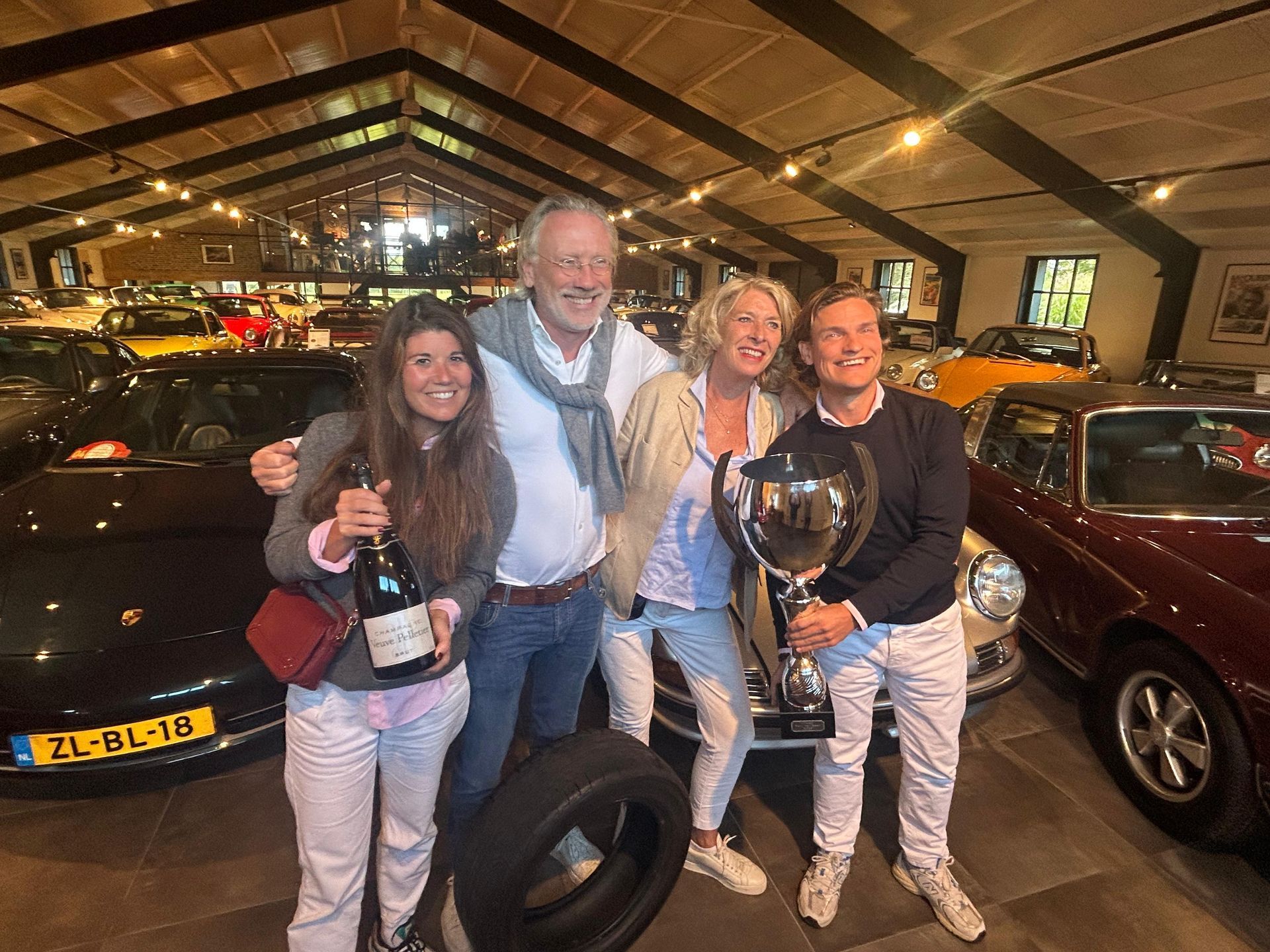 Een groep van vier viert feest in een ruimte met een autocollectie. Een van hen houdt een trofee vast, een ander een fles champagne. Op de achtergrond zijn auto's te zien.