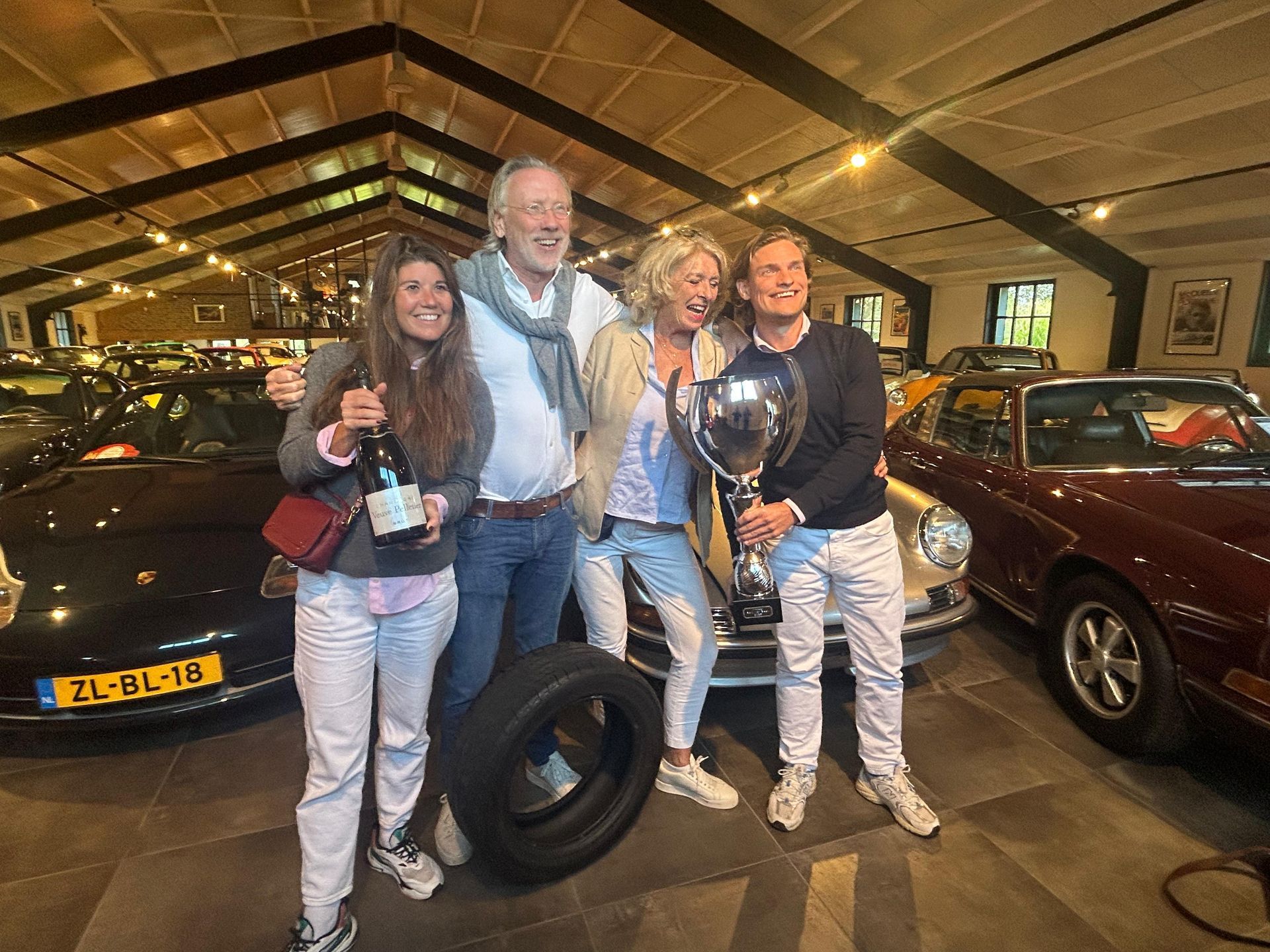 Een groep van vier mensen viert feest bij klassieke auto's in een garage; ze houden een trofee, een band en een fles wijn vast.