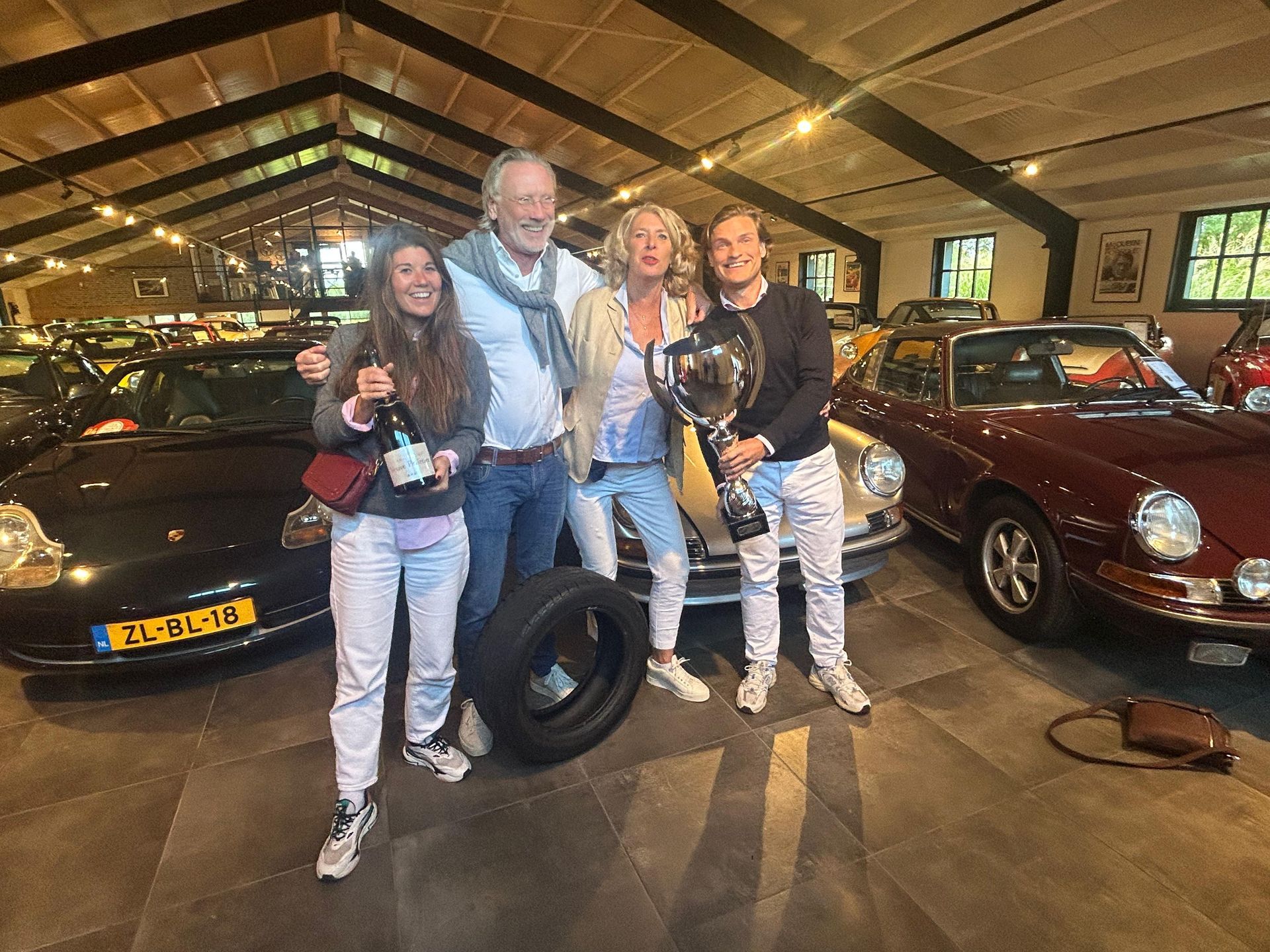 Een groep van vier mensen viert feest bij klassieke auto's. De ene man houdt een trofee vast, de andere een fles champagne.