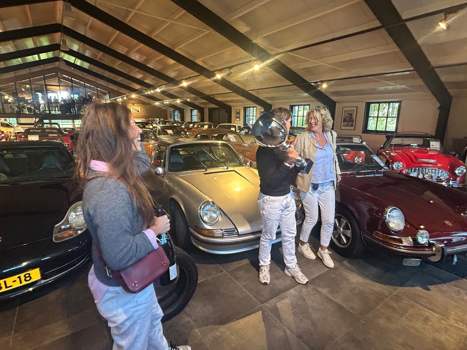Drie vrouwen poseren met klassieke auto's in een showroom. Een van hen draagt een helm.