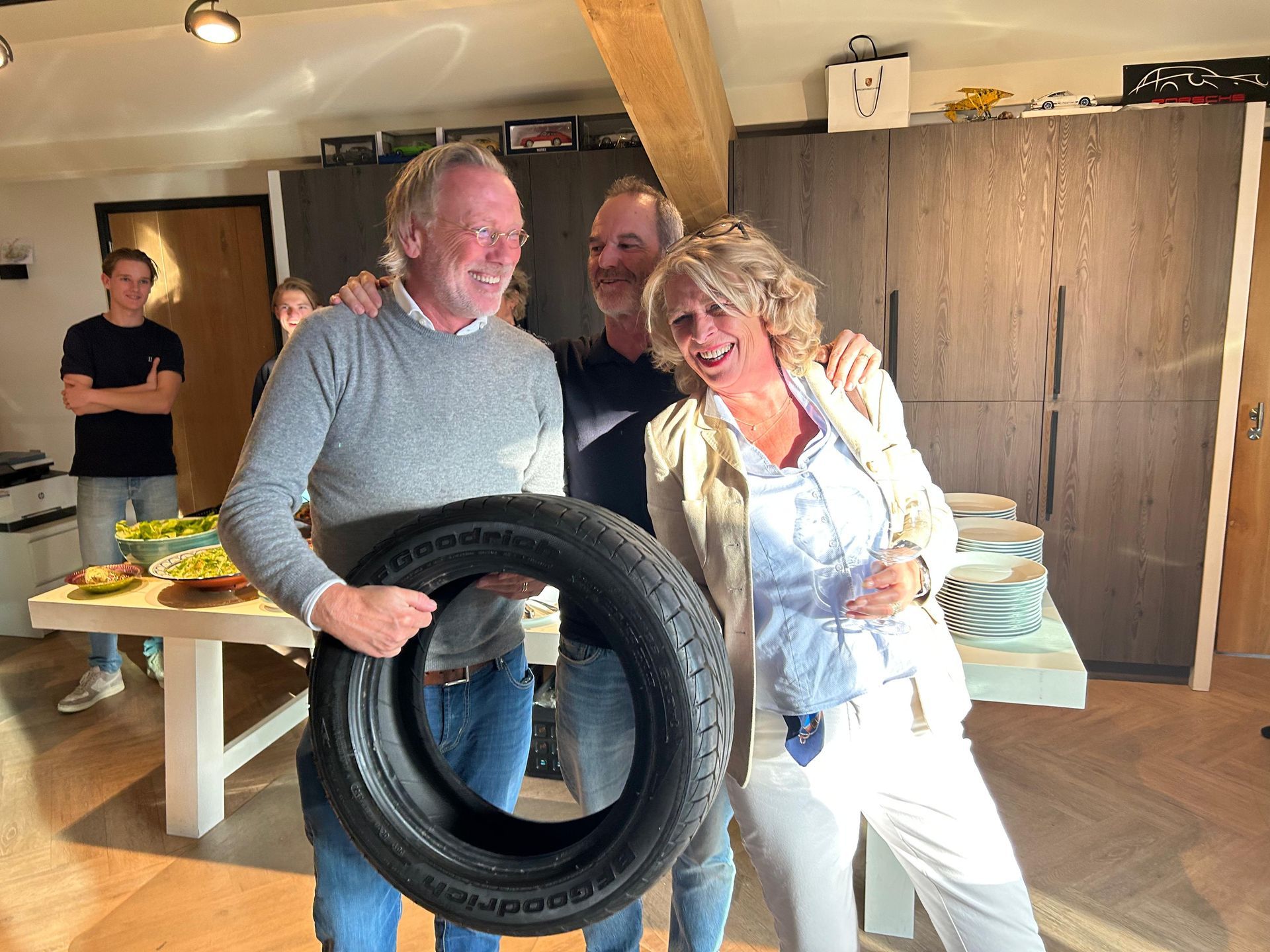 Drie lachende volwassenen met een autoband. De persoon links houdt de band vast, de anderen hebben hun armen om elkaar heen geslagen. Binnen in een huis.