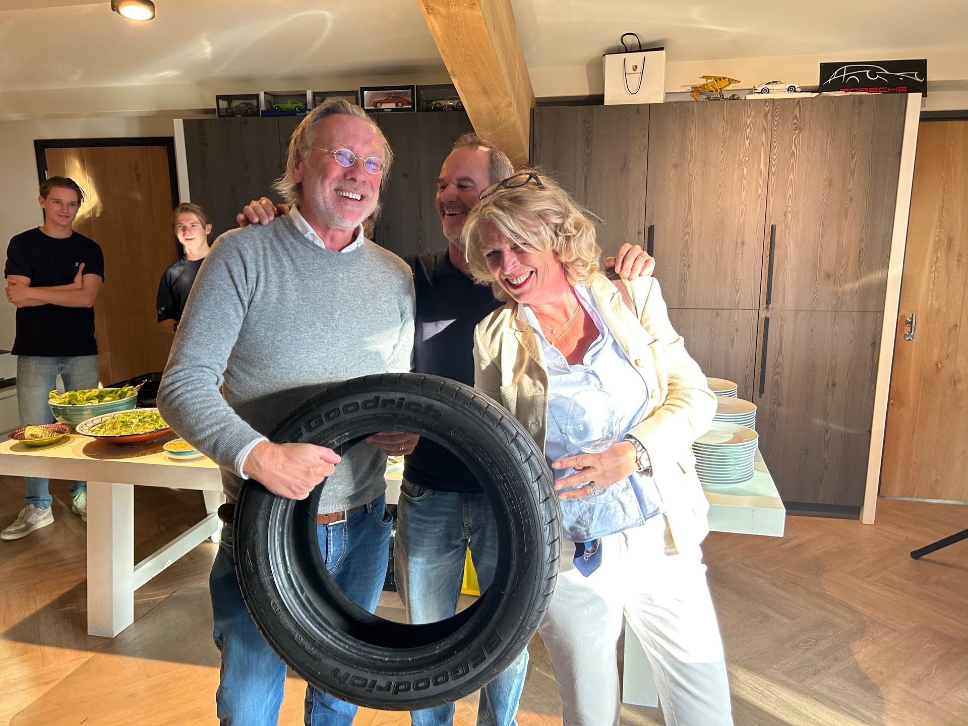 Drie mensen lachen, één van hen houdt een autoband vast. Binnen in een lichte kamer met een tafel, schalen en kasten.
