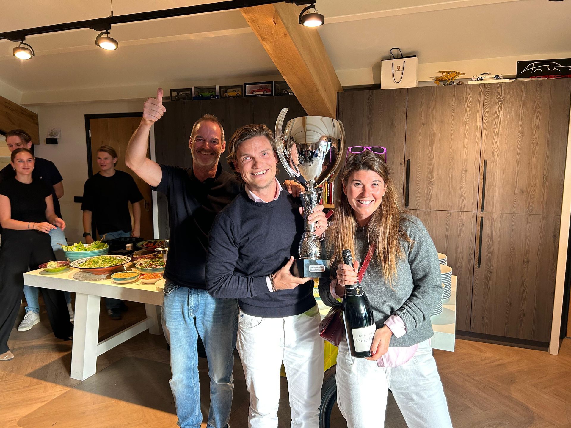 Mensen vieren het binnen met een trofee en champagne; sommigen steken enthousiast hun armen in de lucht.