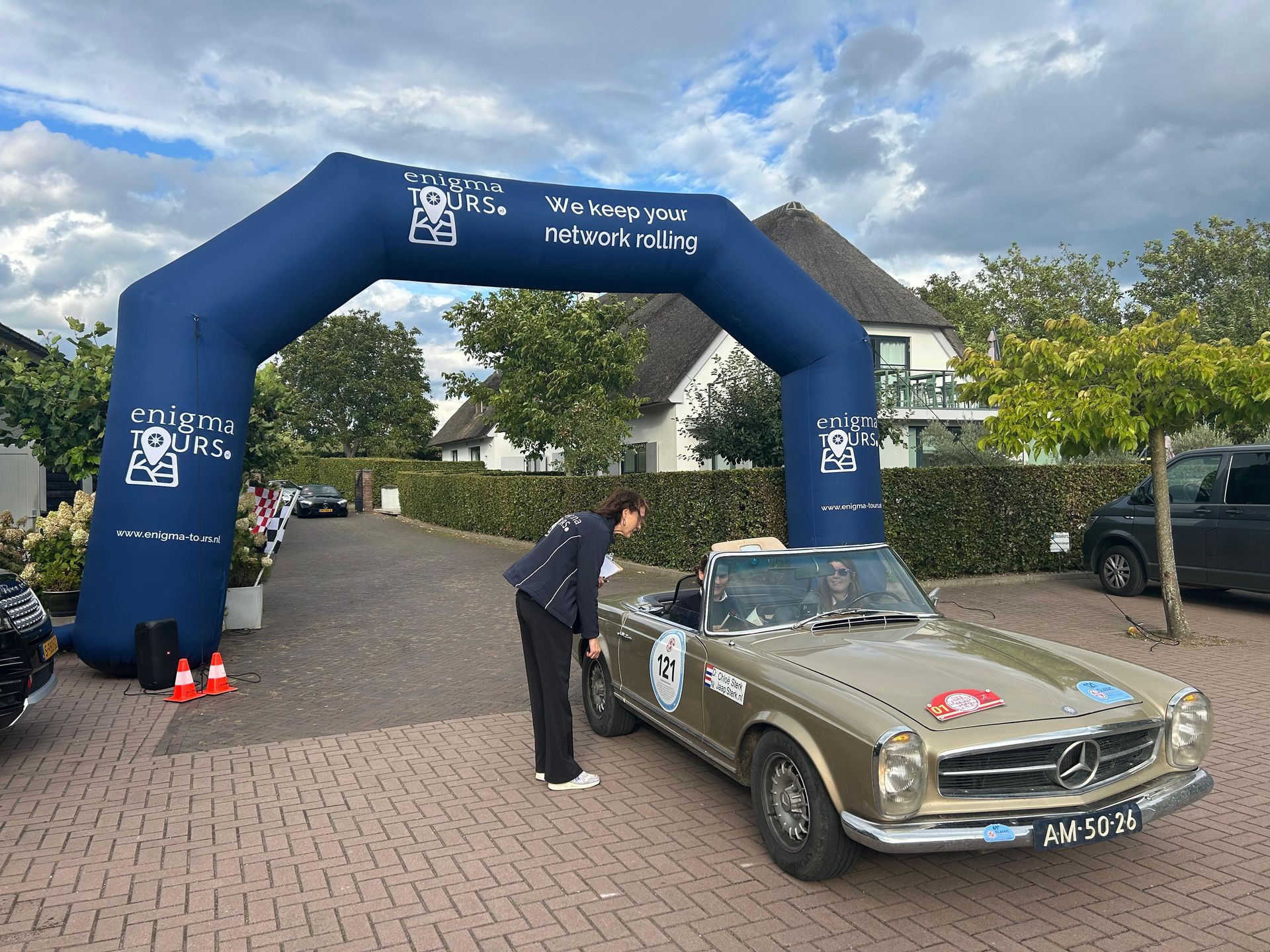 Man in donkere kleding bij een gouden oldtimer onder een blauwe opblaasbare boog.