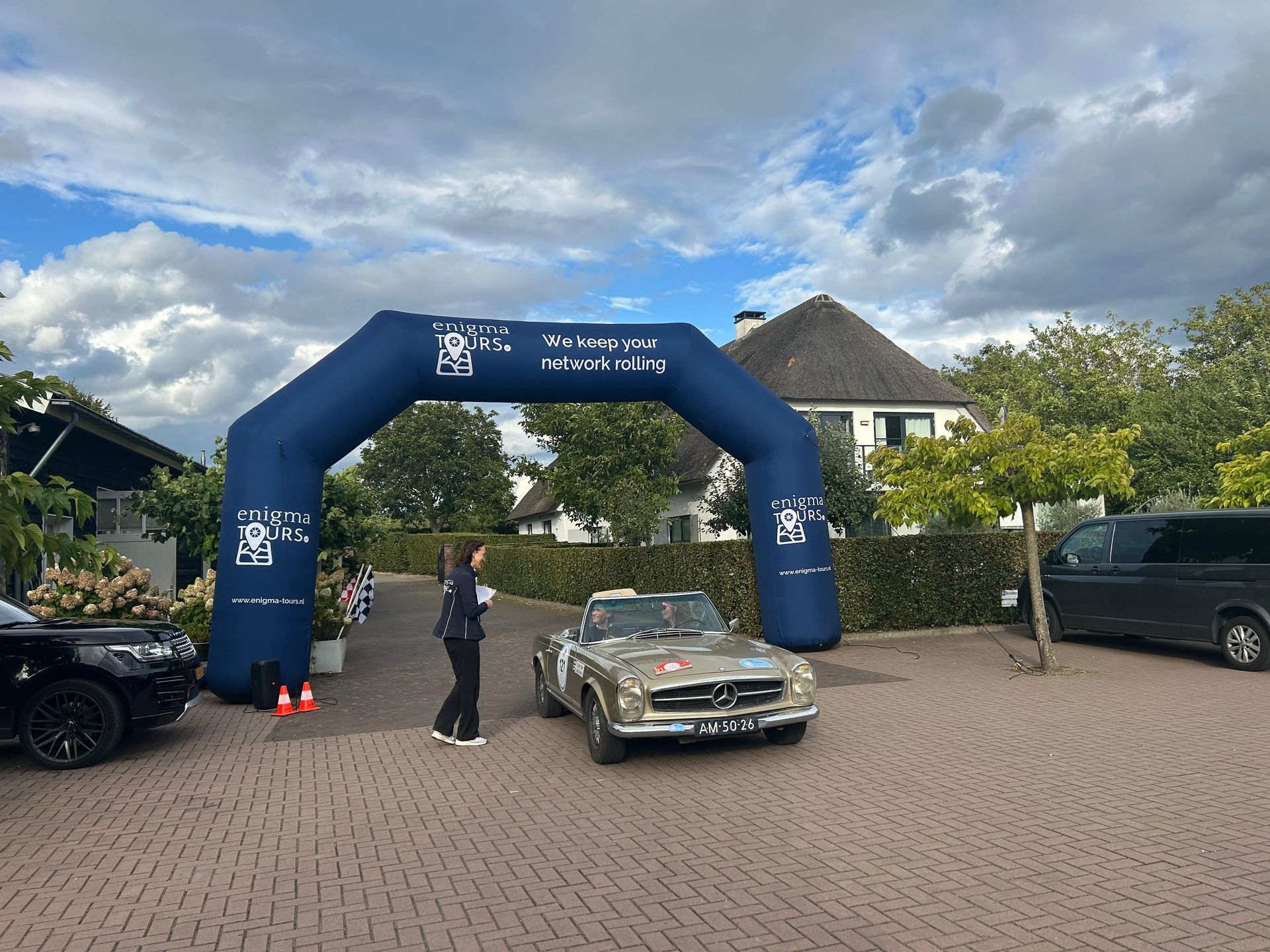 Klassieke auto onder een blauwe boog, evenementbanner, persoon in de buurt, bakstenen oprit.