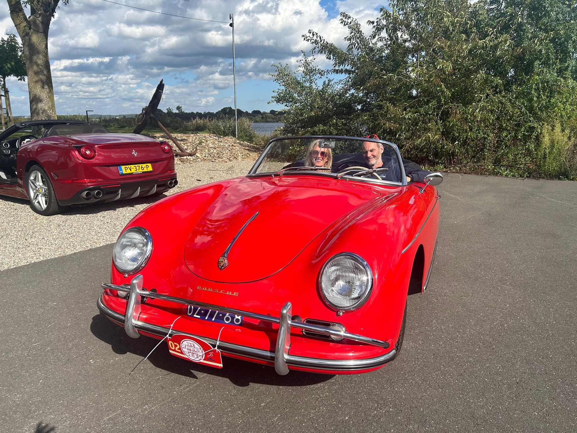 Rode Porsche cabriolet met twee inzittenden, rode Ferrari op de achtergrond. Buitenopname.