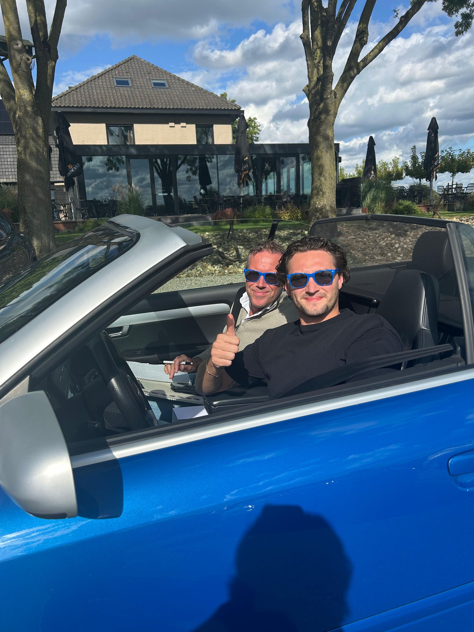 Twee mannen in een blauwe cabriolet, met zonnebrillen op, lachend en met hun duim omhoog. Huis en bomen op de achtergrond.