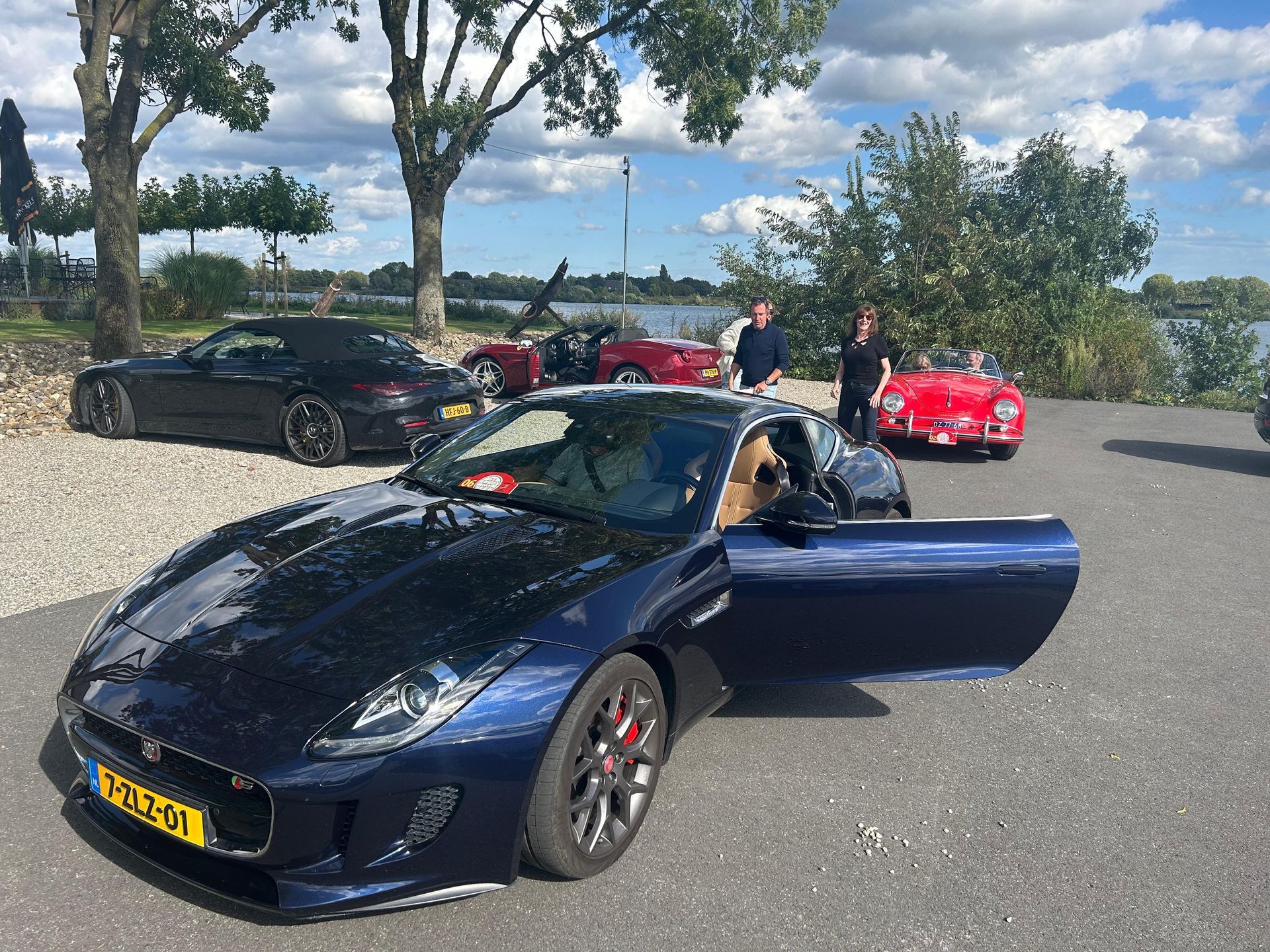 Blauwe Jaguar sportwagen met open deur, geparkeerd tussen andere auto's in de buurt van water en bomen.