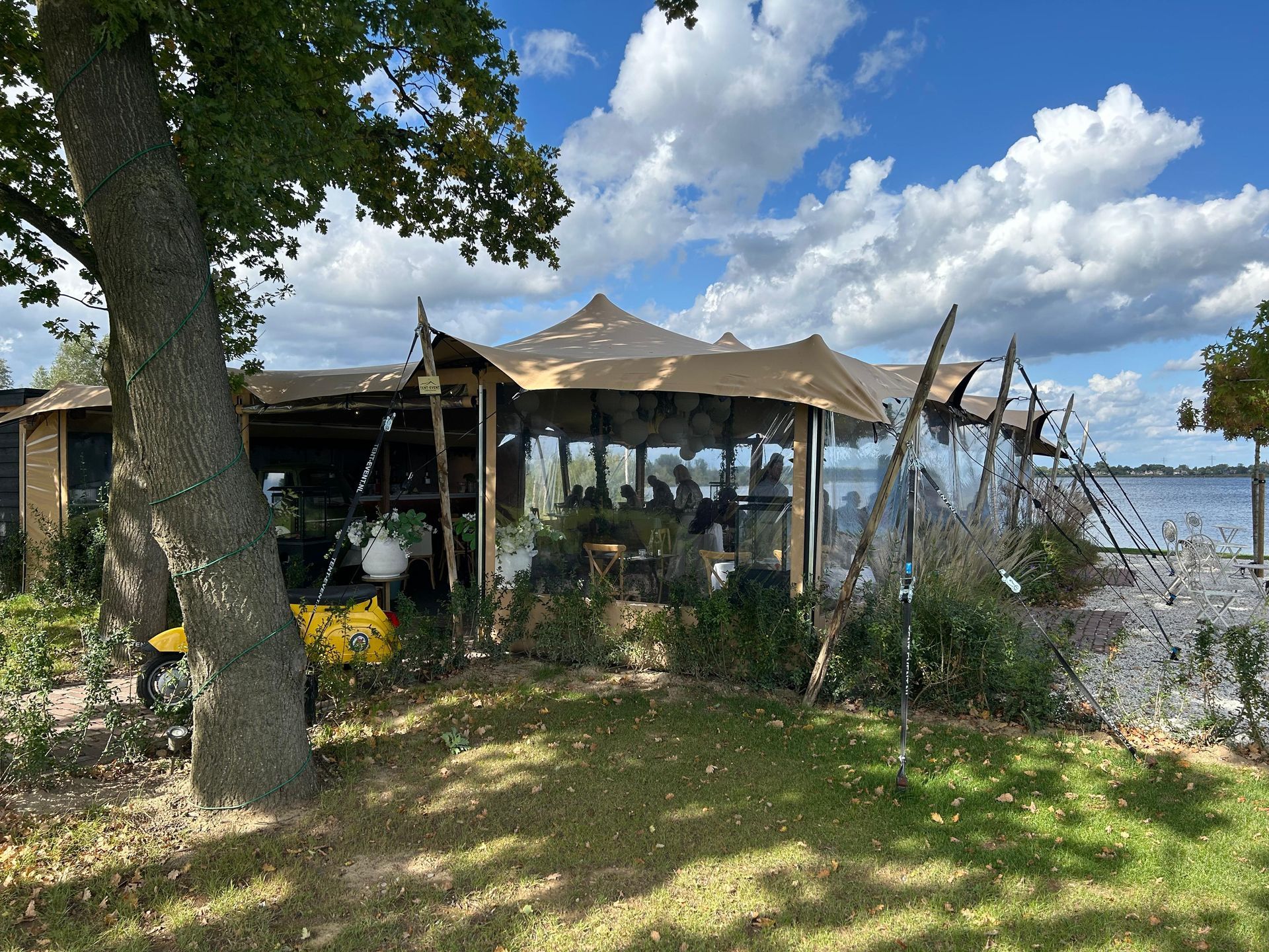 Buitenrestaurant met een tentvormig dak en transparante zijpanelen. Groen gras, een meer en bomen op de achtergrond.